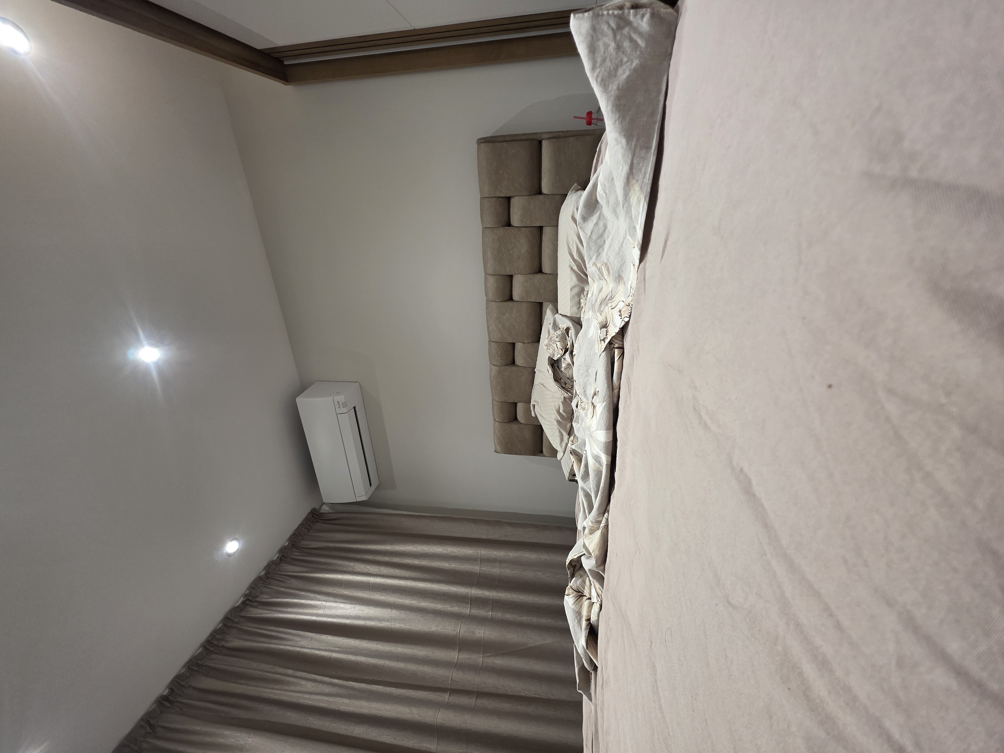 This image captures an interior view of a bedroom in Yagodovo, Bulgaria. The primary subject is a bed, partially visible on the right side of the frame, covered with plain, light-colored bedding. A large, upholstered headboard, constructed from rectangular blocks in a complementary light brown hue, is situated behind the bed.
On the left wall, a white air conditioning unit is mounted high up. Below it, light brown curtains with visible pleats cover the lower portion of the wall, suggesting the presence of a window. The room's ceiling features two bright, recessed spotlights that create noticeable glare, illuminating the space. A decorative wooden beam runs along the top edge of the room where the ceiling meets the wall. The walls are painted a uniform, light, neutral color. No specific activity is taking place, and no people are visible. A small red item, possibly a container or cup, can be faintly seen near the headboard. No text is discernible in the image. The lighting suggests an indoor setting with artificial illumination.