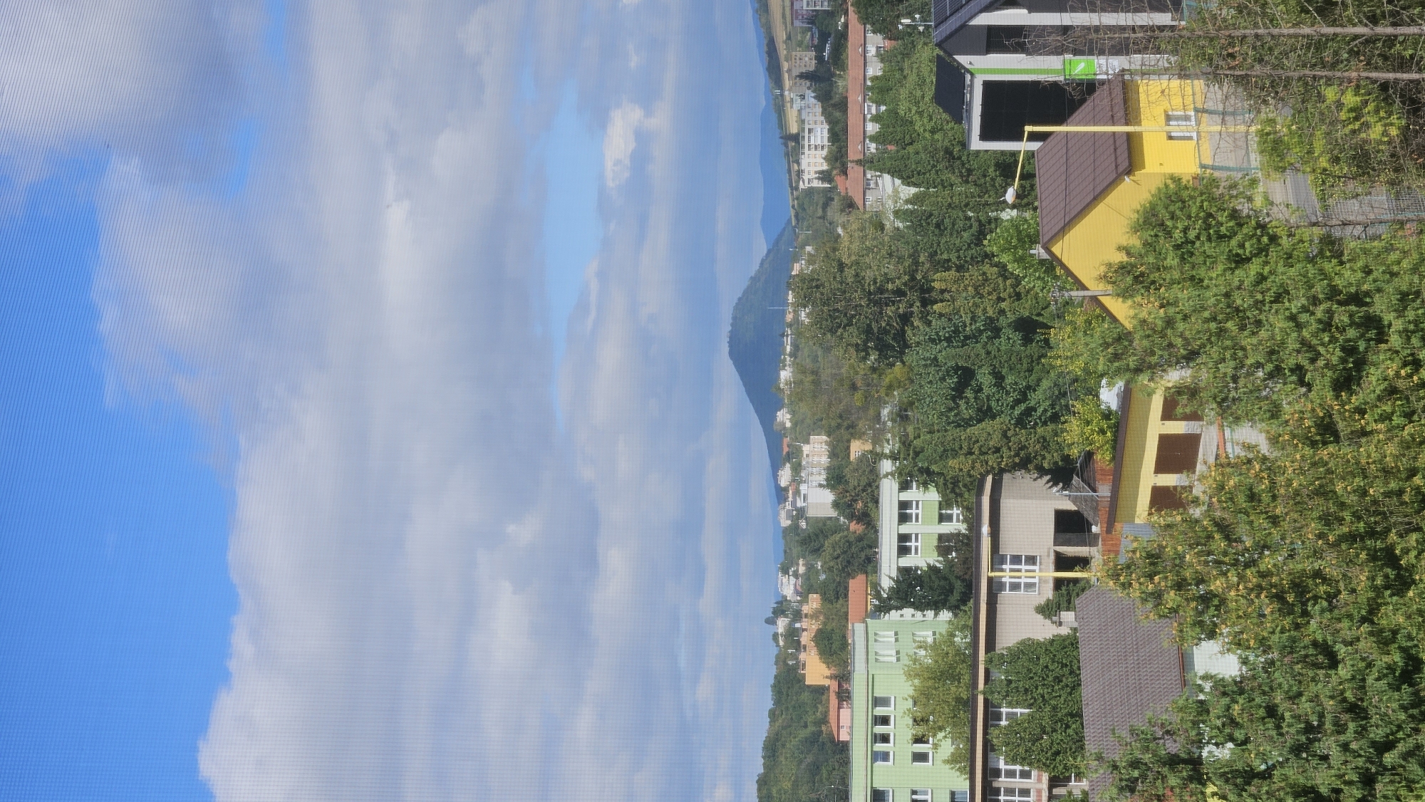 This image depicts a residential area in Prešov, Slovakia, with a prominent conical hill in the background. The foreground is filled with lush green trees and foliage, obscuring some of the lower structures. In the mid-ground, several buildings are visible. A bright green, multi-story building with numerous windows is situated on the left. Adjacent to it, another building of similar height features a grey facade and white-framed windows. Further to the right, a brown-roofed house with a concrete facade and large windows is partially visible. A small, bright yellow house with a brown roof and a single window is prominent in the lower right section, with a tall yellow lamppost beside it. Behind this yellow house, a long, low building with brown garage doors can be seen. The background displays a suburban landscape with various buildings and trees. The most striking feature of the background is a dark, conical hill with a tree-covered slope, typical of the Prešov region. The sky is a vibrant blue with scattered white and grey clouds. The overall impression is a sunny day in a green, hilly urban environment.