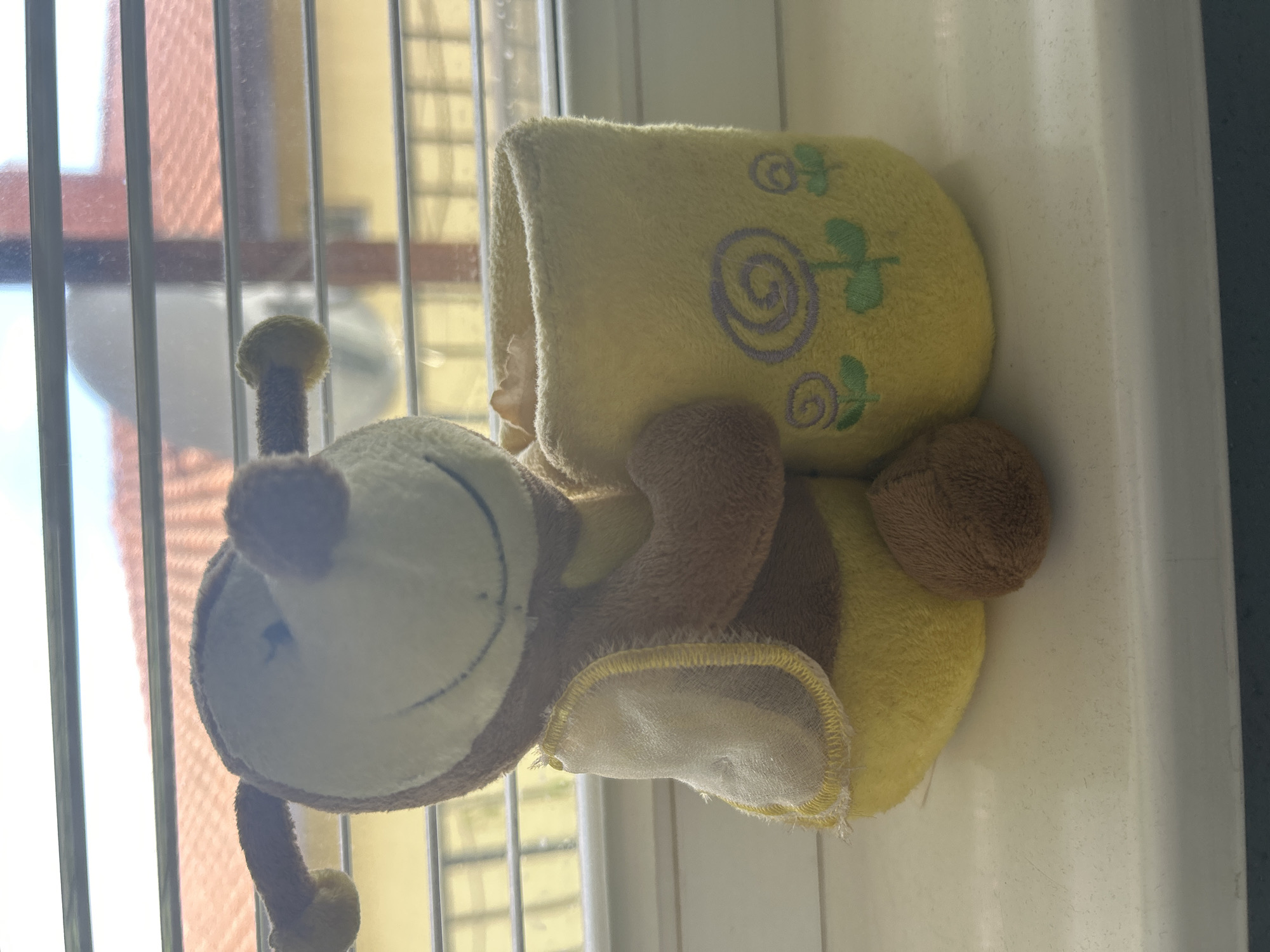 The main subject of the image is a plush toy bee, positioned next to a soft, yellow slipper or baby bootie. The bee toy features a white face with a faint blue stitched smile, brown body sections, yellow stripes, and small antennae with yellow tips. The slipper is made from a fuzzy yellow fabric and is decorated with light purple swirling patterns and green leaf or flower motifs.
Both items are resting on a white, slightly textured surface, which appears to be a windowsill or a shelf. Behind them, a window is visible, characterized by prominent vertical bars, suggesting either window blinds or security grilles. Through the blurred glass of the window, an outdoor scene is discernible, showing parts of a reddish-brown building or wall and other lighter, yellowish structures in the distance, indicating a daytime setting.
The scene is brightly illuminated by natural light, suggesting it is daytime. There is no activity taking place; the items are static. There is no visible text on any of the subjects. The location context provided is Dunavtsi, Bulgaria, though no specific visual cues in the image itself explicitly confirm this, other than the general appearance of the background architecture which could be typical for the region.