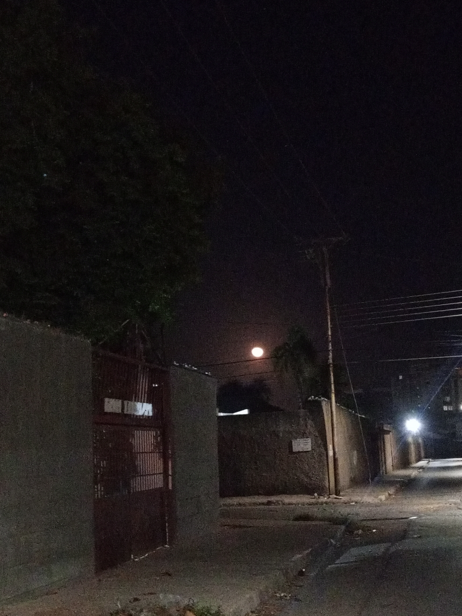 A dark, low-angle shot shows a street scene in Porlamar, Venezuela at night. The main elements are a tall concrete wall with a red metal gate on the left, a telephone pole with wires stretching across the frame, and the street receding into the distance. A few dimly lit buildings and streetlights are visible down the street. A full moon is visible in the sky above the wall and telephone pole. There are no visible people or interactions. The scene appears quiet and desolate.