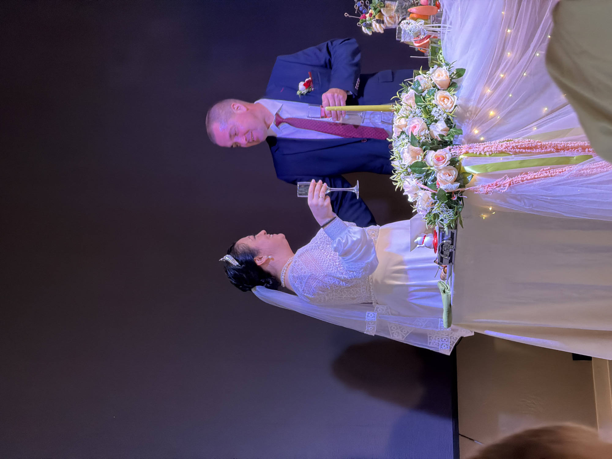 A wedding reception is taking place in Domodedovo, Russia. A bride wearing a white lace dress and a tiara is toasting with a champagne flute. She is facing a groom in a dark blue suit and red tie, who is also holding a champagne flute. They are standing behind a table decorated with flowers, sheer fabric, and fairy lights. The background is a dark wall.