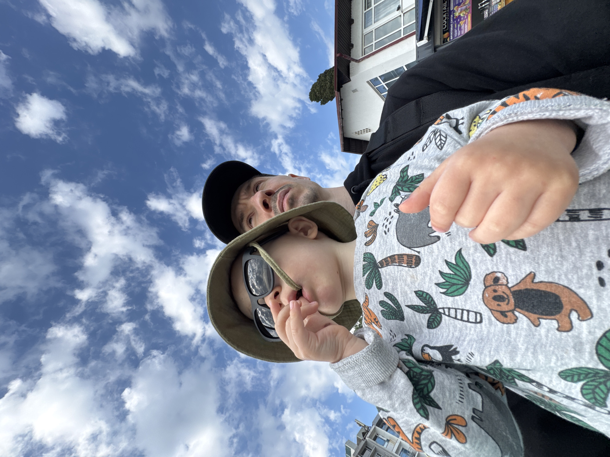 A low-angle, upward-tilted shot captures a man and a young child against a backdrop of a bright blue sky dotted with scattered white clouds. The man, wearing a black baseball cap, is positioned behind and slightly above the child. The child is the focal point, dressed in a light gray patterned shirt and a green bucket hat, also sporting dark sunglasses. The child is holding their fingers near their mouth. The scene includes architectural elements of buildings, including a white building with a red roof and windows. There is also an advertisement poster for cocktails and GOKU visible. The location is possibly in Sochi, Russia.