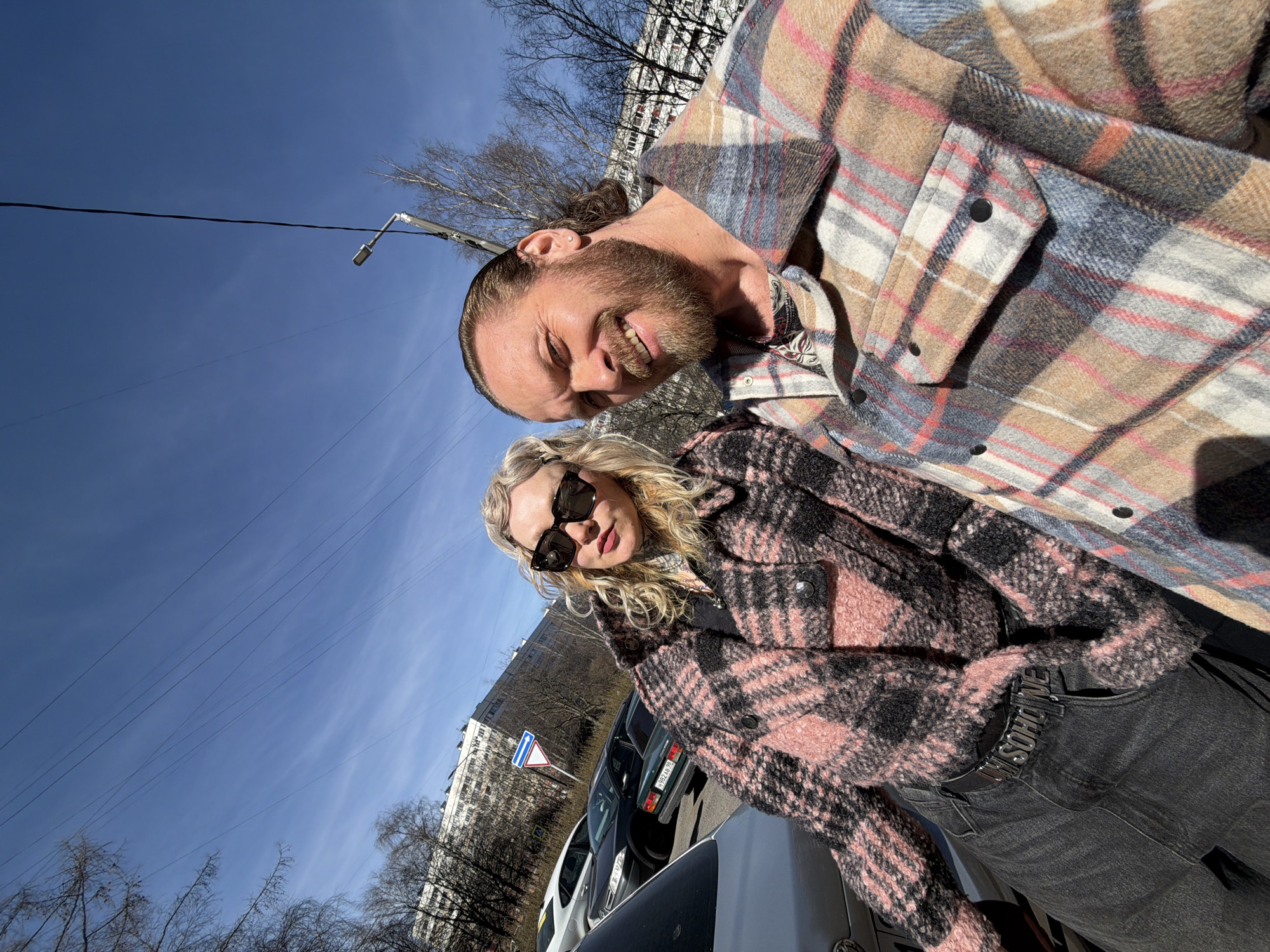 A sunny day in Saint Petersburg, Russia, presents a selfie of two people standing near parked cars. The man, sporting a beard and long hair pulled back, wears a plaid shirt. The woman has blonde curly hair and is dressed in a pink and black plaid coat with sunglasses. The background features a building and a road sign. Overhead, the sky is clear and blue with bare trees and electrical wires visible.
