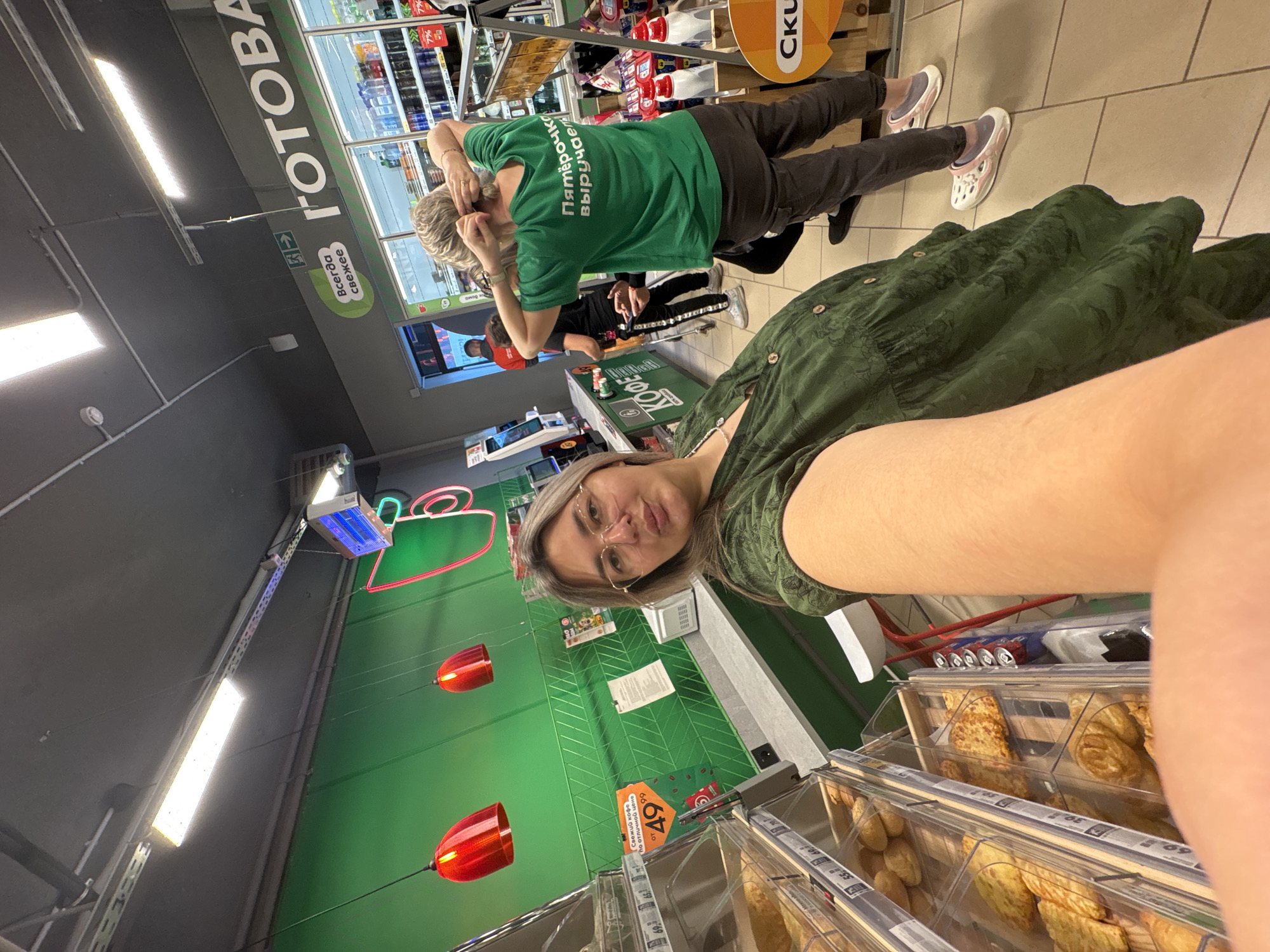 The image shows a woman in a green dress taking a selfie in a grocery store. She is wearing glasses and has blonde hair. In the background, there are shelves of baked goods and drinks. Another woman in a green t-shirt with Russian writing on the back is visible, as well as a few other people and store displays. The overall environment suggests a typical supermarket setting. The specific location in Birobidzhan, Russia, cannot be determined from the image alone.