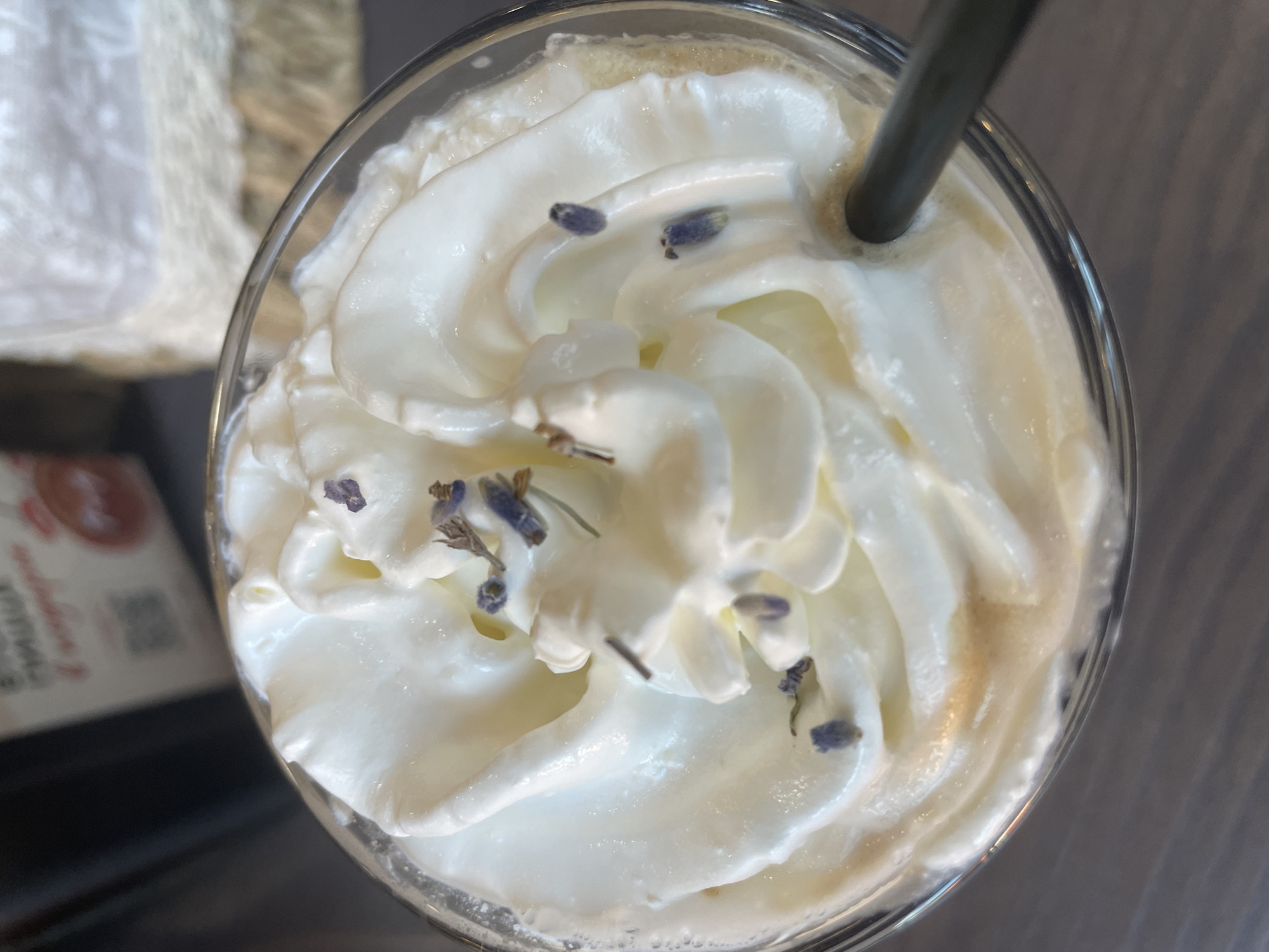 A close-up, top-down shot shows a glass filled with a creamy white beverage, topped with whipped cream and garnished with a few sprigs of dried lavender. A dark straw is partially submerged in the drink. The background is blurred, but appears to be an indoor setting, possibly a cafe, with some out-of-focus signage and decorative elements. The image is captured in Kaliningrad, Russia.