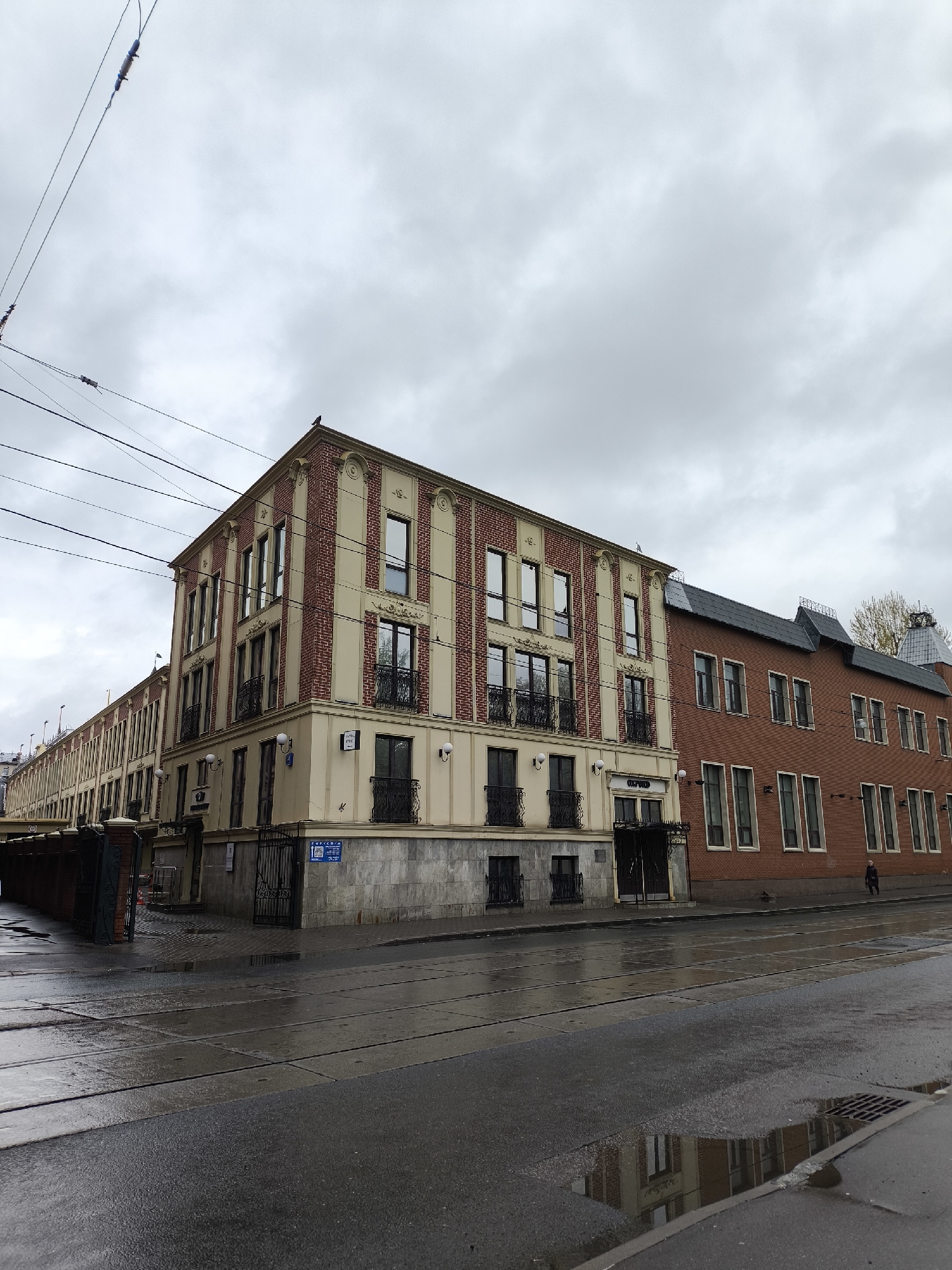 The image displays an urban street scene in Moscow, Russia, under an overcast sky, with the street visibly wet from recent rain. Two buildings define the street corner, casting reflections on the water-slicked asphalt.
The building on the left features a distinctive facade composed of alternating vertical sections of reddish-brown brick and light cream-colored stucco or stone. It has multiple floors with rectangular windows, some adorned with decorative black iron balconies. The ground level is clad in darker gray stone, with a dark metal gate visible at the corner. A small, dark bird perches on the highest point of its roofline. Overhead utility wires crisscross the sky above this building.
Adjacent to it, on the right, stands a building constructed primarily of reddish-brown brick, featuring numerous tall, narrow windows and a dark, sloped roof. A single individual, appearing to be a man, is walking on the sidewalk in front of this brick building in the distance.
The street itself is dark asphalt, marked by visible seams or lines, possibly indicating old tram tracks. A prominent puddle in the foreground reflects the building facades. A drain grate is visible in the bottom right corner. Visible text on a blue sign at the corner of the cream-and-brick building reads "ТАРУСА" (TARUSA), along with the number "19" and a QR code. The word "OXFORD" is also partially visible on the facade of the cream-and-brick building above an entrance. The overall atmosphere is quiet and subdued due to the cloudy weather.