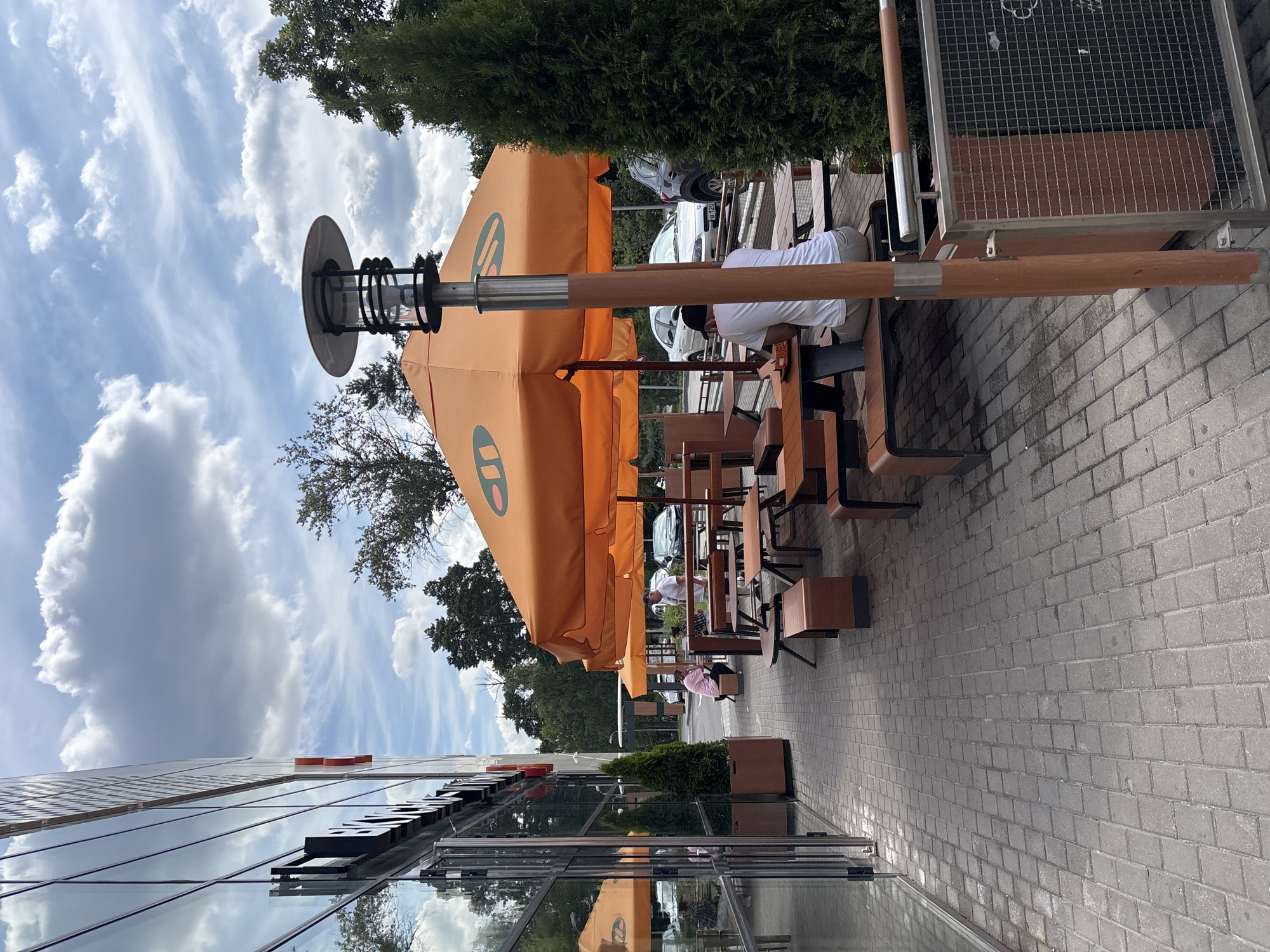 The image depicts an outdoor seating area of a fast-food establishment, likely a McDonald's due to the prominent orange umbrellas featuring the brand's logo. The scene is set on a paved walkway adjacent to a modern building with large glass windows. Several wooden picnic tables and benches are arranged under the umbrellas, with some patrons seated. In the background, cars are parked in a lot, and trees form a natural border around the area. The sky is cloudy, suggesting it might be a partly sunny day. The location is identified as Gorki-2, Russia.