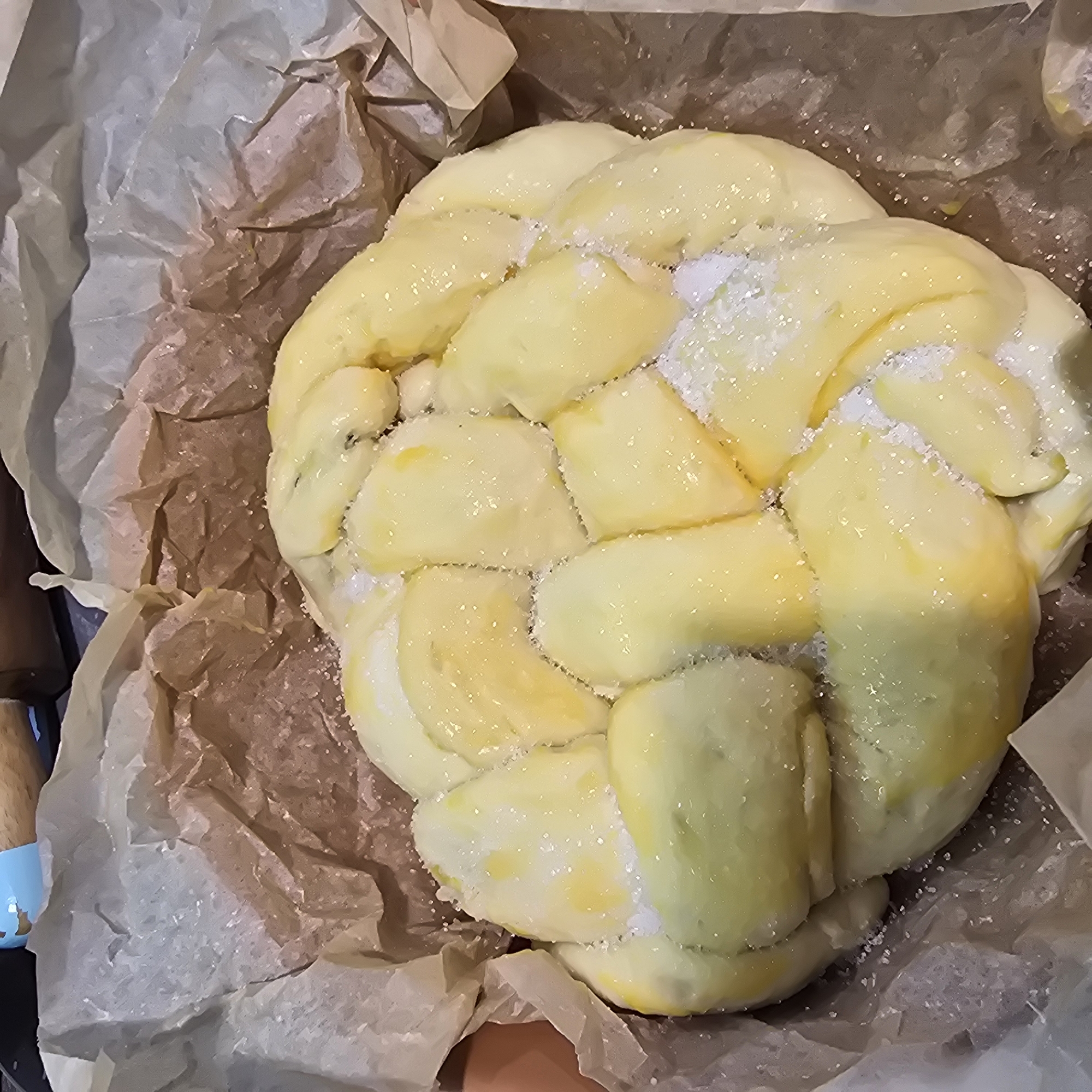 A close-up, top-down view shows a braided, round loaf of dough coated with granulated sugar on a sheet of crumpled parchment paper. The dough appears pale yellow and slightly shiny, suggesting it has been brushed with an egg wash or similar before being sprinkled with sugar. The parchment paper underneath is mostly brown, with some areas appearing lighter and more translucent. Part of a rolling pin with a blue handle is visible in the lower left corner, suggesting the bread has been prepared in a kitchen setting. There's no indication in the image that would pinpoint this in Plovdiv, Bulgaria.