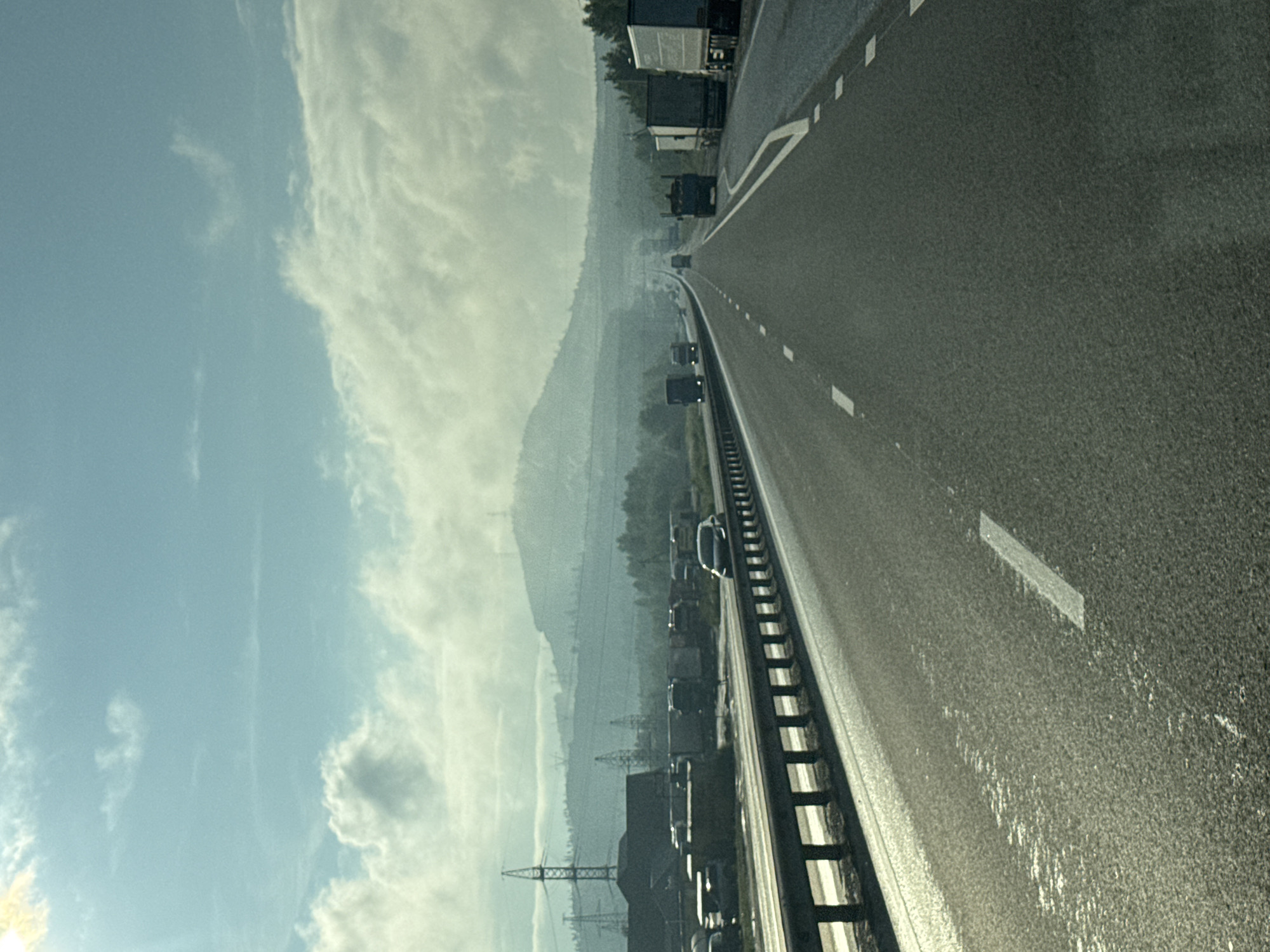 An outdoor, eye-level, vertical shot captures a highway in Revda, Russia, under a hazy, overcast sky. The sun is partially visible in the upper left, casting a faint glare. The highway stretches into the distance, lined with guardrails and marked by white dashed lines. Several semi-trailer trucks and cars are traveling on the road. In the background, a range of hills with scattered trees and industrial structures, including power lines and pylons, can be seen through the atmospheric haze. The landscape suggests a somewhat industrial or developing area.