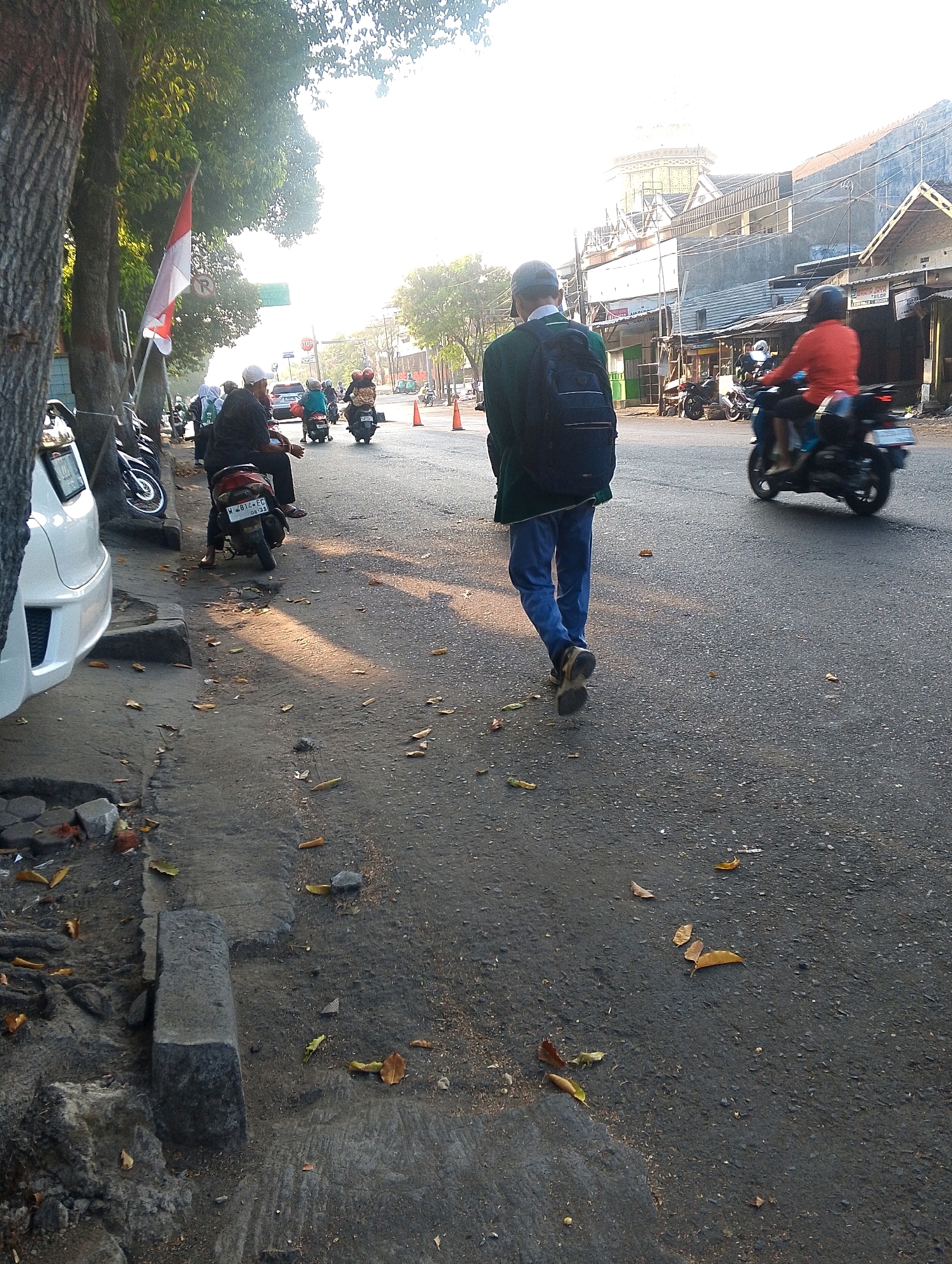 A man wearing a green jacket and blue pants, carrying a black backpack, walks away from the camera on the right side of a street. The sun is setting or rising, casting long shadows. In the background, other people ride motorcycles, and buildings line the street. A white car is parked on the left side of the image. The street is paved with asphalt and there are fallen leaves on the ground. A white and red flag is visible on the left side of the image.