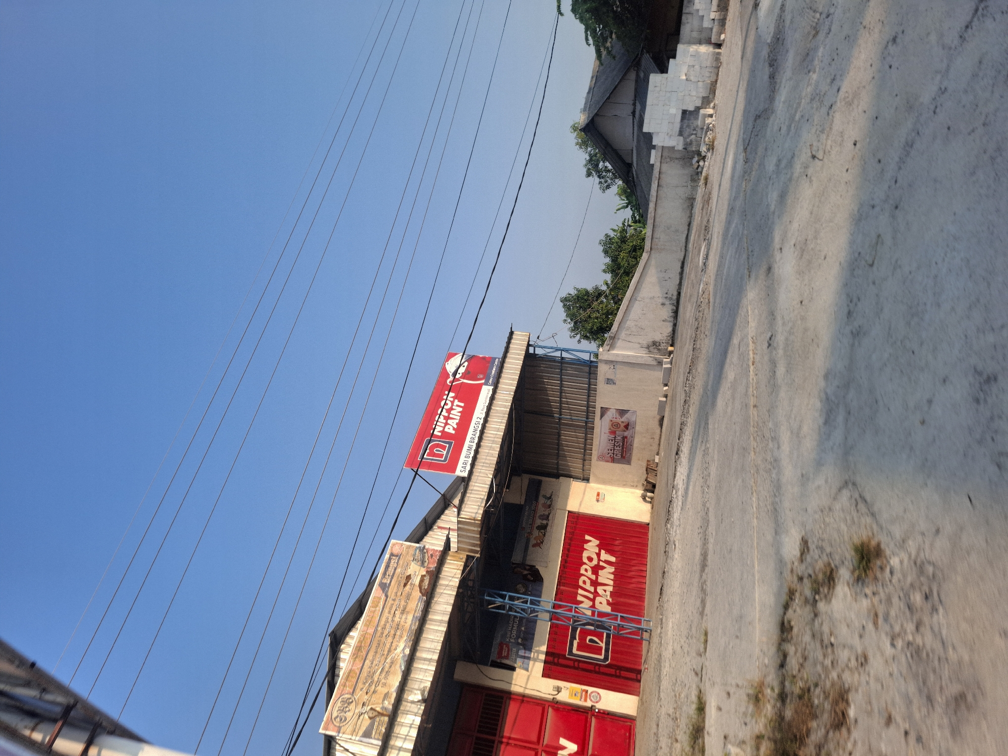 A wide-angle, eye-level shot captures an outdoor scene in Sumberwudi, Indonesia, on a clear, sunny day. The image is dominated by a bright blue sky crisscrossed by several power lines. In the midground, a commercial building with a red and white facade is prominent. The building features a large red corrugated metal door with the "NIPPON PAINT" logo. Above, a red and white sign also displays "NIPPON PAINT" and text that appears to be an address or company name. To the left of the main entrance, a smaller red door is visible.
The building is adjacent to a concrete lot, which shows signs of wear and tear, with patches of dry vegetation. A white concrete wall runs along the perimeter of the property, separating it from the surrounding area. Beyond the wall, rooftops of other buildings and lush green trees are visible. To the right of the main building, a stack of white building blocks rests against the wall. The overall impression is one of a commercial area in a tropical climate.