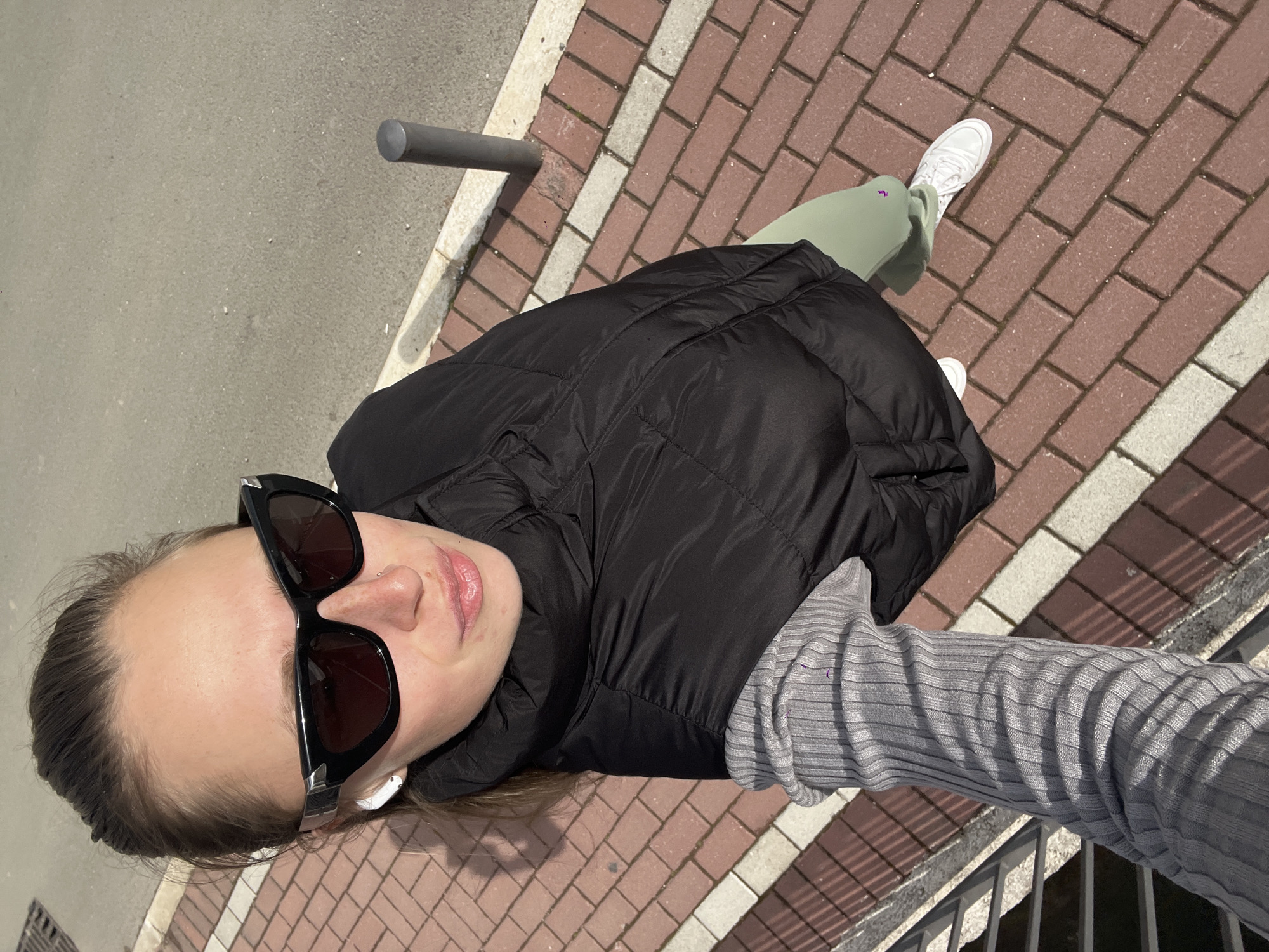 A person, presumably female, is taking a selfie outdoors on a paved area with brick-like tiles. They are wearing sunglasses, a black puffer jacket, a grey long-sleeved shirt, light green pants, and white sneakers. The paved area is a combination of red and white/grey bricks. In the background, there is a grey post and a grey concrete surface. To the right, there is a grey railing. The location is likely Rafailovići, Montenegro, although this is difficult to ascertain definitively from the image alone. The person is holding out their arm to take the selfie.