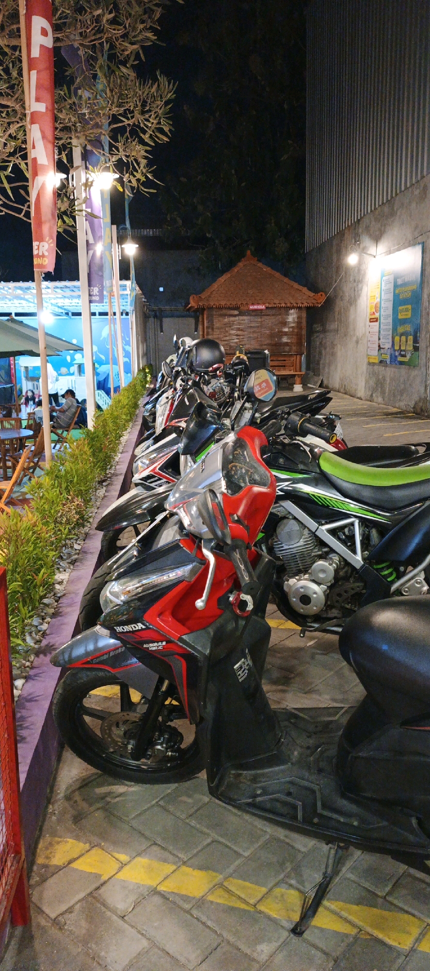 The image captures a nighttime scene in what appears to be an outdoor seating area and motorcycle parking lot. In the foreground, a row of parked motorcycles is visible, the closest one being a red and black scooter with the "HONDA" logo clearly displayed. The motorcycles are parked on a textured gray pavement marked with yellow lines. To the left, a red metal barrier encloses a landscaped area with green bushes and some scattered rocks. Beyond the bushes, an outdoor seating area with wooden tables and chairs is set up under umbrellas. People are seated at the tables, dimly lit. In the background, a small traditional Balinese-style wooden structure with a thatched roof stands between the seating area and the motorcycles. Further in the background, a textured concrete wall rises, with a large poster or sign attached to it. The overall lighting suggests it is after sunset, with artificial lights illuminating the area. The presence of motorcycles and outdoor seating suggests this may be a restaurant, cafe, or similar establishment. The location is identified as Sidoarum, Indonesia.