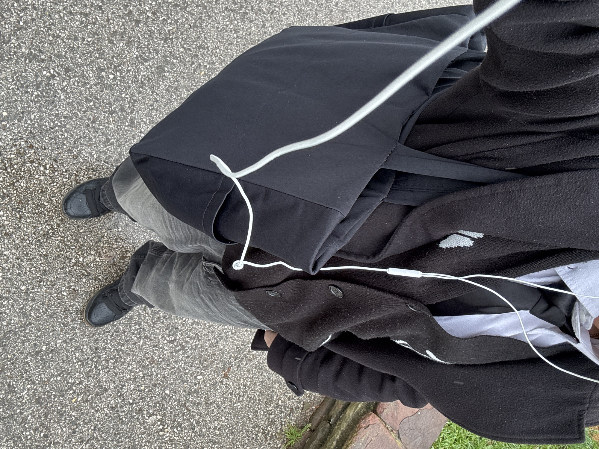 The image depicts a person standing on an asphalt surface, likely a sidewalk or street. The person is wearing gray jeans, black sneakers, and a black jacket over a white shirt. They also have a black tote bag slung over their shoulder and white headphones plugged into their ears. The background shows a portion of a brick border or wall, and some green grass. The location is assumed to be the city of Sofia, Bulgaria.