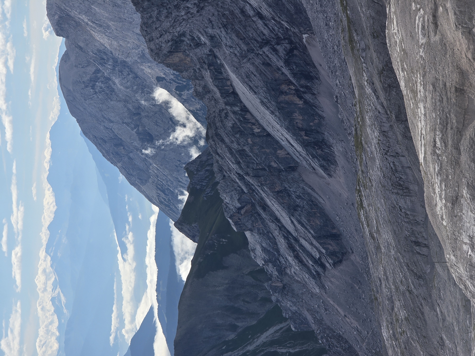 This is a vertical, medium shot of a rugged mountain landscape with layers of peaks receding into the distance. The foreground is a rocky, barren slope. In the middle ground, a steep, dark gray mountain face is deeply fissured and shows evidence of scree slopes. Further back, a lighter gray mountain peak rises, with some wisps of white clouds clinging to its sides. In the background, successive layers of blue mountains fade into a hazy sky dotted with white clouds. The scene is likely a view from a high vantage point in the Bavarian Alps, possibly near Garmisch-Partenkirchen, Germany, given the characteristic dramatic peaks and valleys of the region. There are no people or discernible man-made objects in the image.