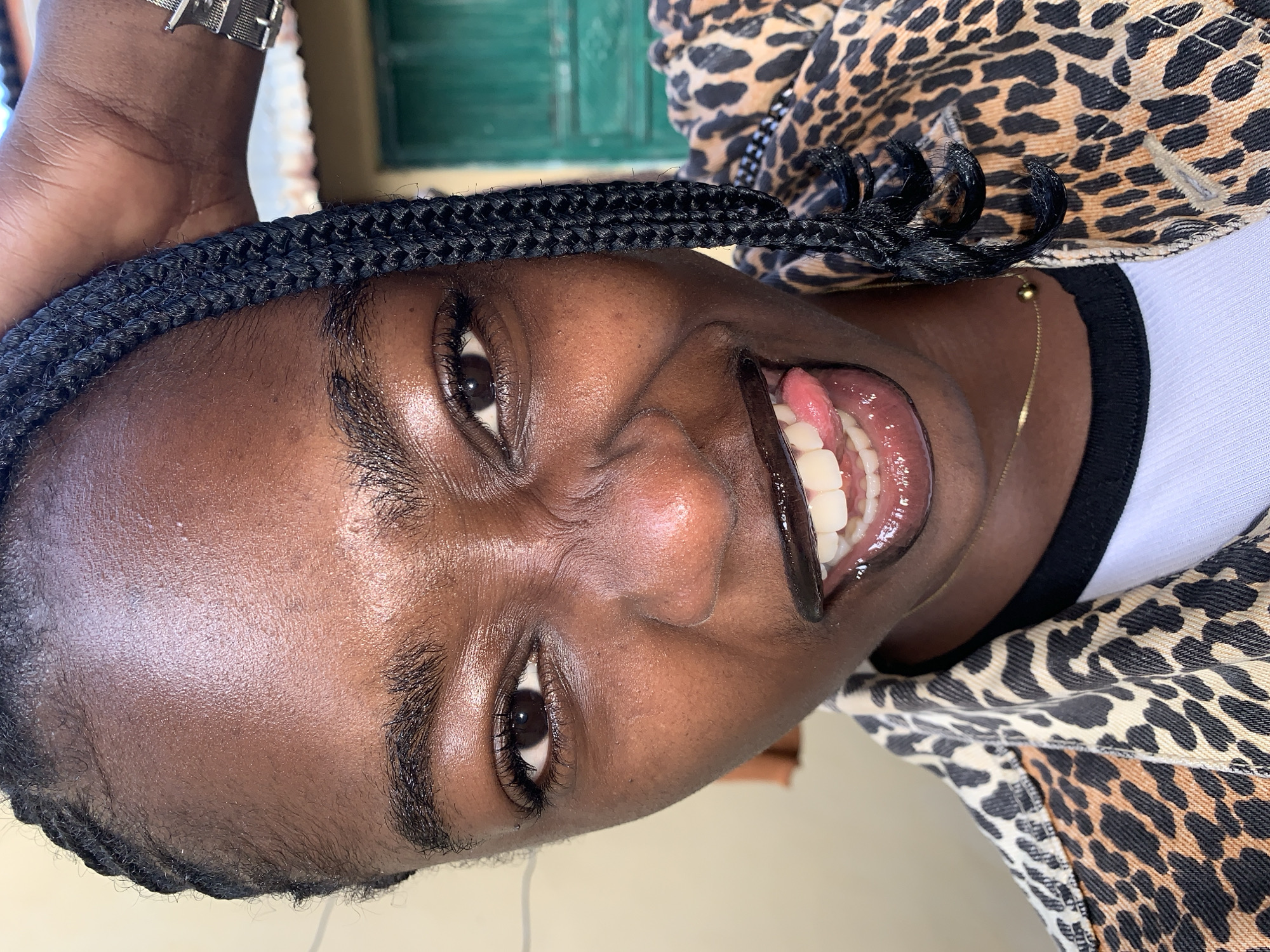 The image is a close-up, frontal, eye-level shot of a young Black woman in Rafin Par, Nigeria. She has dark skin and her hair is styled in black braids, one of which is draped over her right shoulder. She is wearing a leopard-print jacket over a white t-shirt, and a thin gold necklace with a small bead. Her mouth is open, and she is sticking out her tongue, which is pink. She has dark lipstick on, and her teeth are white. Her eyebrows are dark and arched. Her eyes are dark brown, and her lashes are long. The background is slightly out of focus and appears to be an outdoor setting with a cream-colored wall on the left and a green door or window on the right. The lighting suggests it is daytime.