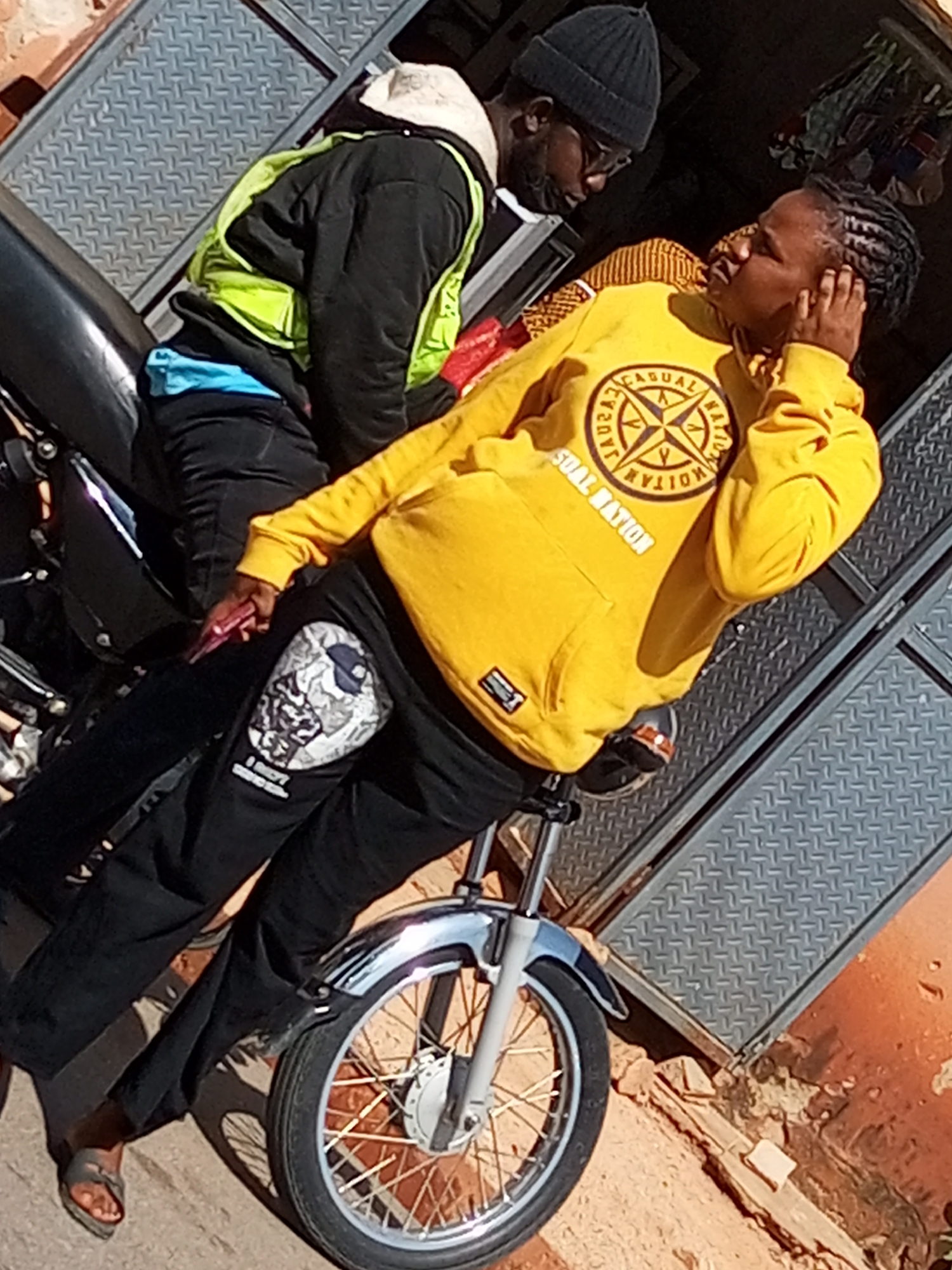 Two individuals are present, one wearing a yellow hooded sweatshirt with a circular emblem on the chest, black pants with a graphic on the left thigh, and grey sandals. The other individual is wearing a dark beanie, glasses, a black jacket over a bright green vest, and blue shorts. They are positioned on what appears to be a motorcycle, with the motorcycle's front wheel and fender visible. The background consists of a corrugated metal structure with a diamond plate pattern. The scene is outdoors, with a reddish-orange wall and ground surface suggesting an urban environment. The image seems to be taken in Gombe, Nigeria.