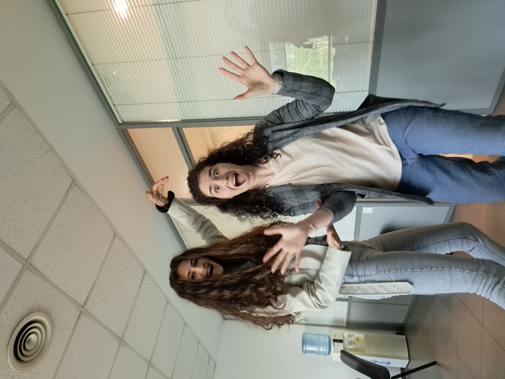 Two young women are posing for a picture indoors. One woman, positioned on the upper right, wears a gray and white checkered blazer over a beige shirt and blue jeans. She has dark, curly hair and an open-mouthed expression with her hands raised. The other woman, located on the lower left, wears a light-colored jacket and light-colored jeans. She also has long, curly, dark hair and an open-mouthed expression with her hands raised.
The setting appears to be an office or institutional environment. The background includes window blinds, a white ceiling with a circular air vent, a wall, and a water dispenser. Given the context, the picture may have been taken in a building in Sofia, Bulgaria, but there's no direct evidence of the city itself in the image.