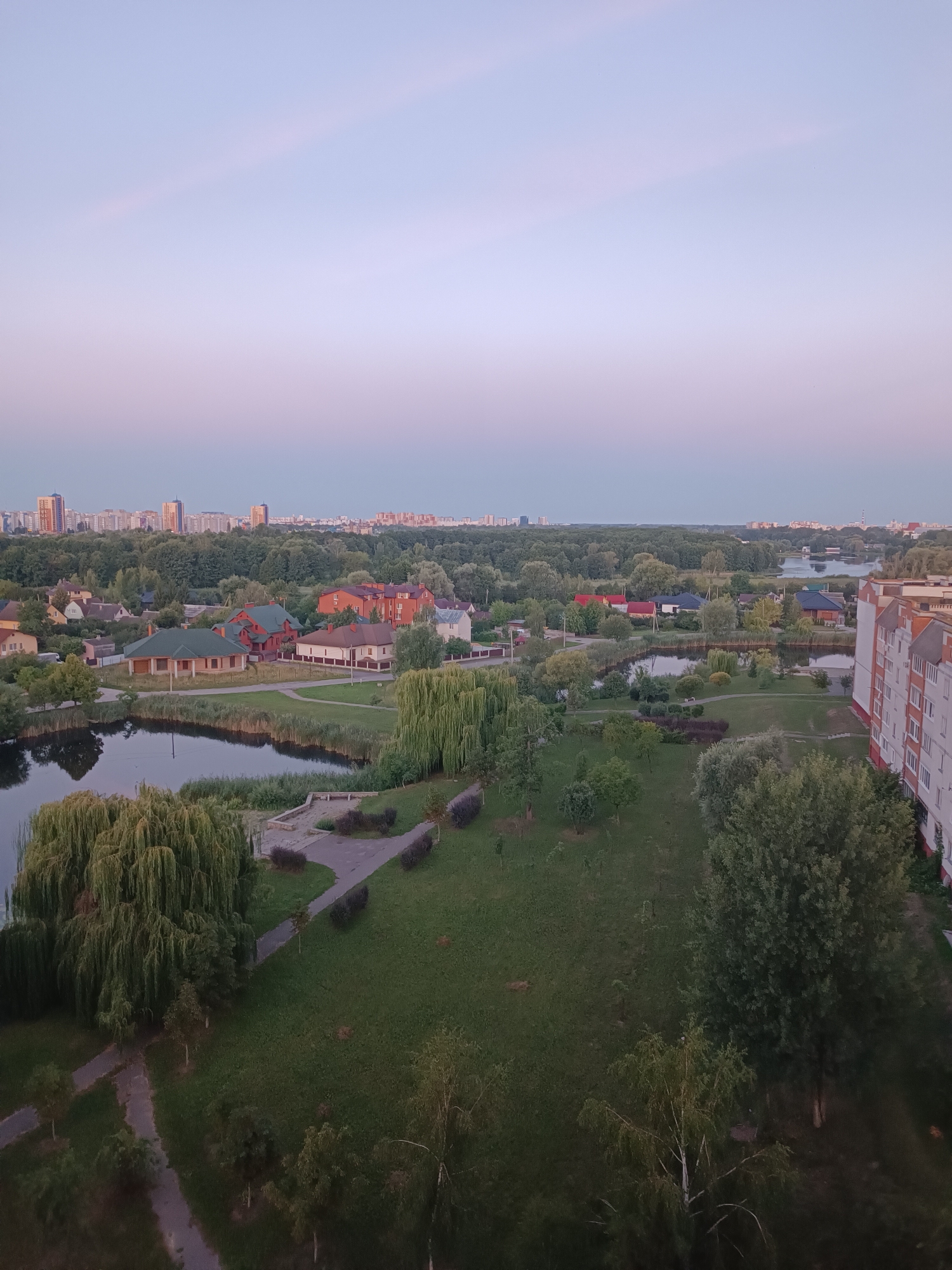 This is an aerial view of a suburban landscape at dusk, likely in Brest, Belarus. The scene is dominated by a twilight sky transitioning from a pale lavender near the horizon to a soft, light blue overhead. In the mid-ground, a small lake or pond is visible, bordered by reeds and weeping willow trees. Beyond the water, a cluster of houses with red and beige roofs are nestled amongst lush green trees. Further in the distance, a cityscape of apartment buildings rises against the horizon. The foreground features a well-maintained park with manicured lawns, paved pathways, and scattered trees. The overall impression is one of tranquility and the quiet transition from day to night. There are no people or visible text in the image.