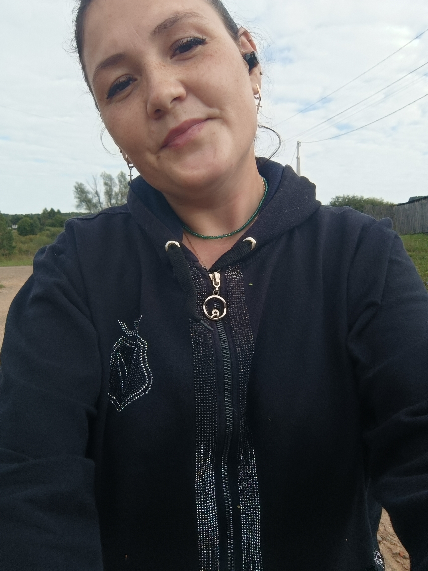 In this medium-shot, head-and-shoulders selfie, a woman with dark hair is outdoors. She is wearing a black hooded jacket with a zipper. The jacket has decorative beadwork on the left chest and along the zipper. She is also wearing a green beaded necklace and cross-shaped earrings. There is a black earbud in her right ear. Her face is turned slightly to the left, and she is looking towards the camera. The background shows a rural landscape with trees, a fence, and utility lines against a cloudy sky.