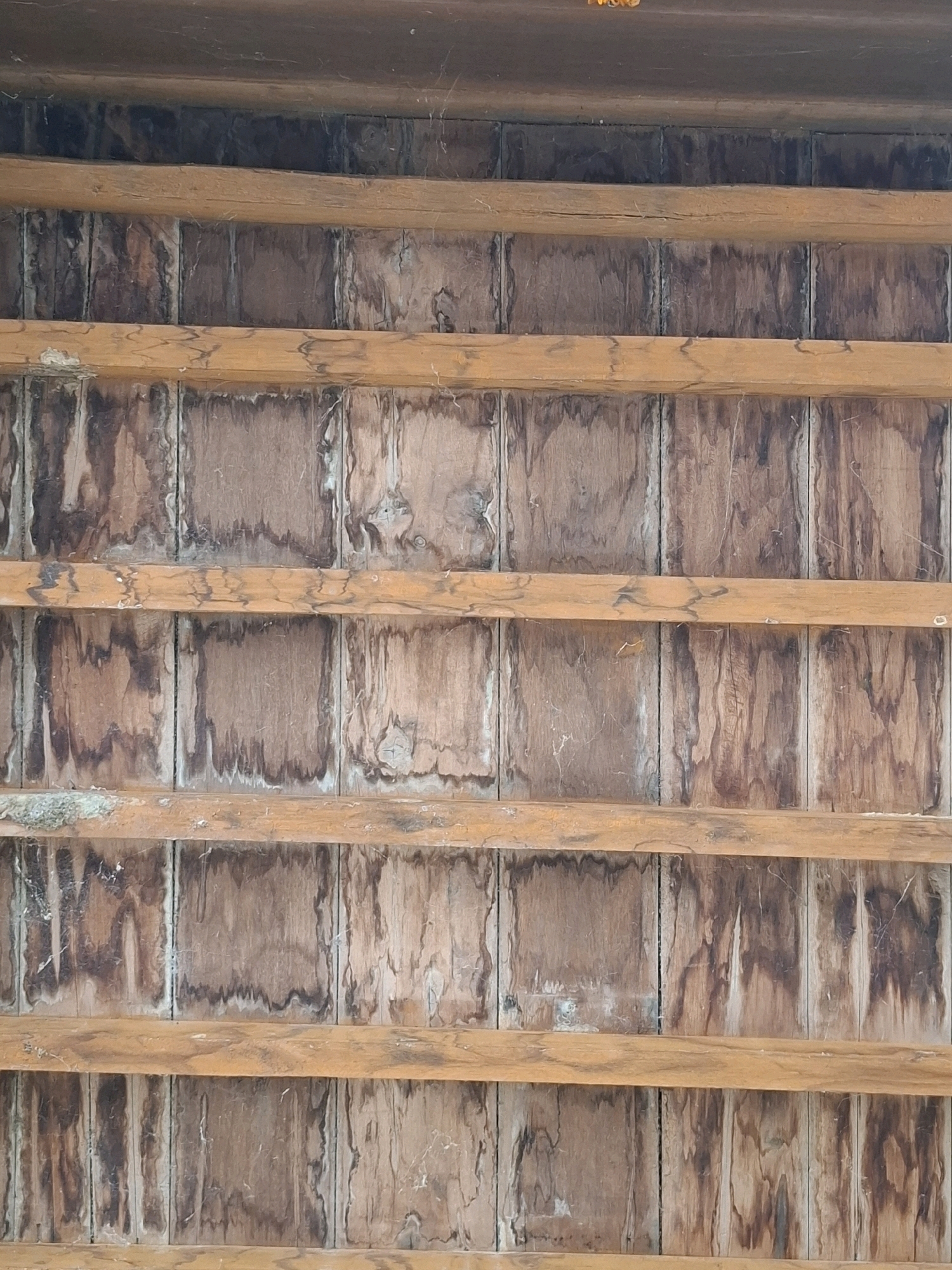 The image depicts a close-up, angled view of a wooden ceiling structure, likely part of an outdoor or unfinished building in Sukon Peudaya, Indonesia. The ceiling is constructed from horizontal wooden beams interspersed with a backing of what appears to be plywood or particle board, divided into vertical sections. The wood exhibits signs of age and weathering, with discoloration, water stains, and a rough texture. Several cobwebs are visible, suggesting a lack of recent maintenance. The overall impression is one of an aged, weathered, and possibly neglected wooden structure.