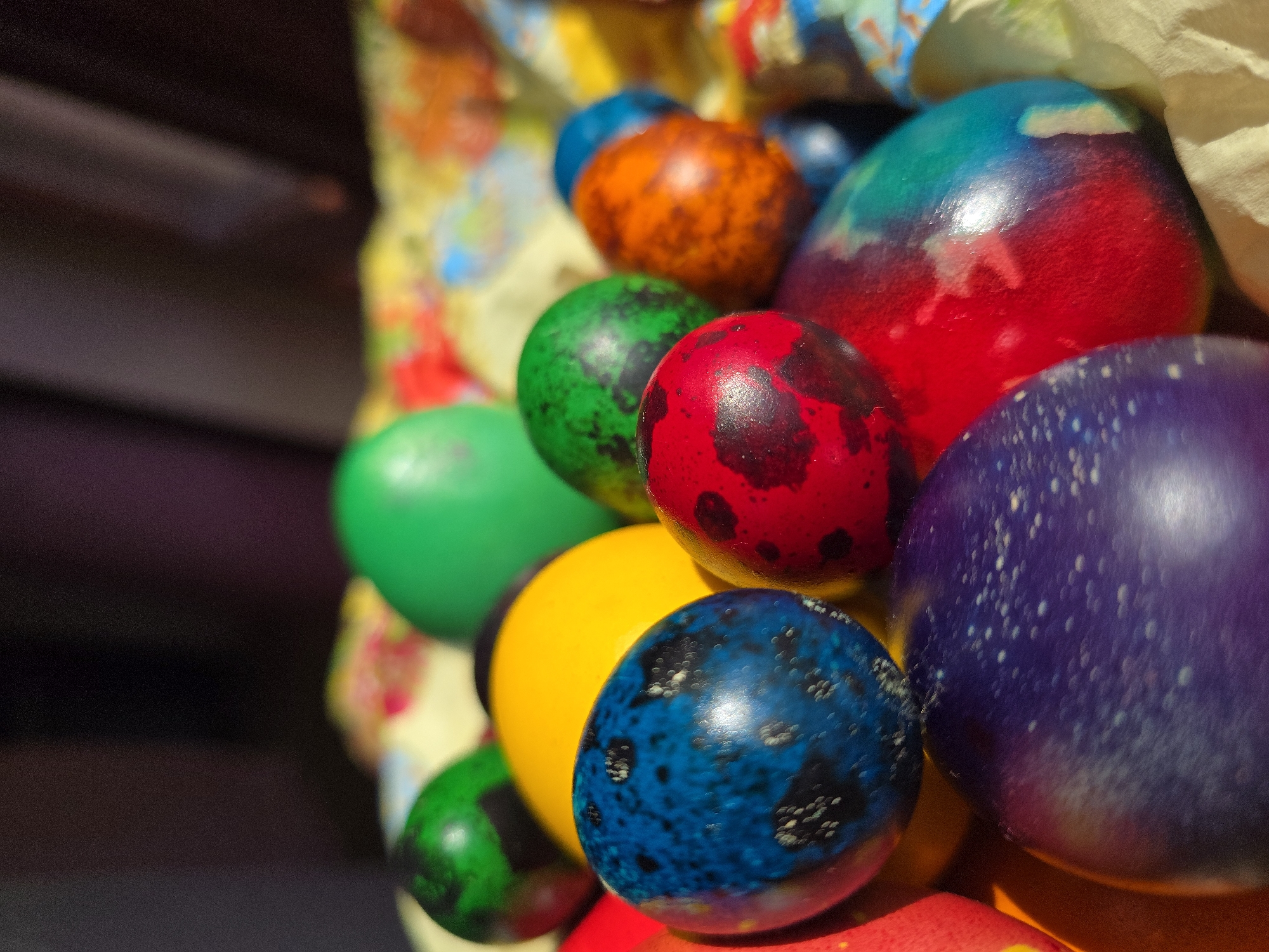 A close-up photograph shows a pile of brightly colored Easter eggs. The eggs are dyed in a variety of colors, including red, blue, green, yellow, orange, and purple. Some eggs are solid colors, while others have spots or tie-dye patterns. The eggs are piled together, filling the frame. The background is out of focus and consists of floral paperboard. The image suggests a festive Easter celebration but contains no information to identify a place such as Varna, Bulgaria.