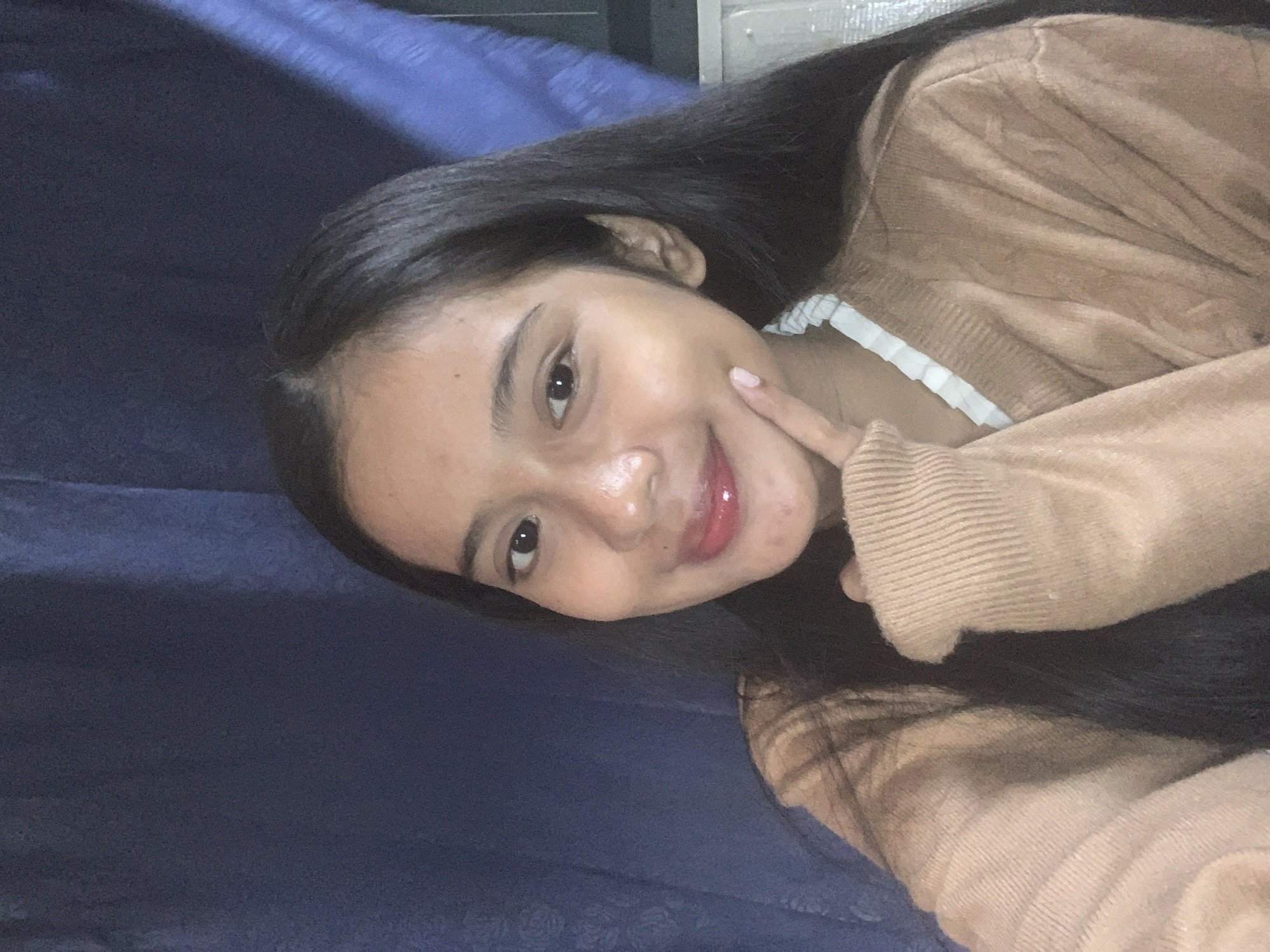 A young person with long, dark hair is taking a selfie indoors. They are wearing a tan cardigan over a white ruffled top. Their left hand is resting on their cheek, and they are looking directly at the camera with a slight smile. The background consists of dark blue curtains and a white object that could be a piece of furniture or an appliance. The lighting suggests an indoor environment with a light source from the front. The image is framed from the chest up. No specific location in Sungai Meriam, Indonesia is identifiable from the visual information provided.