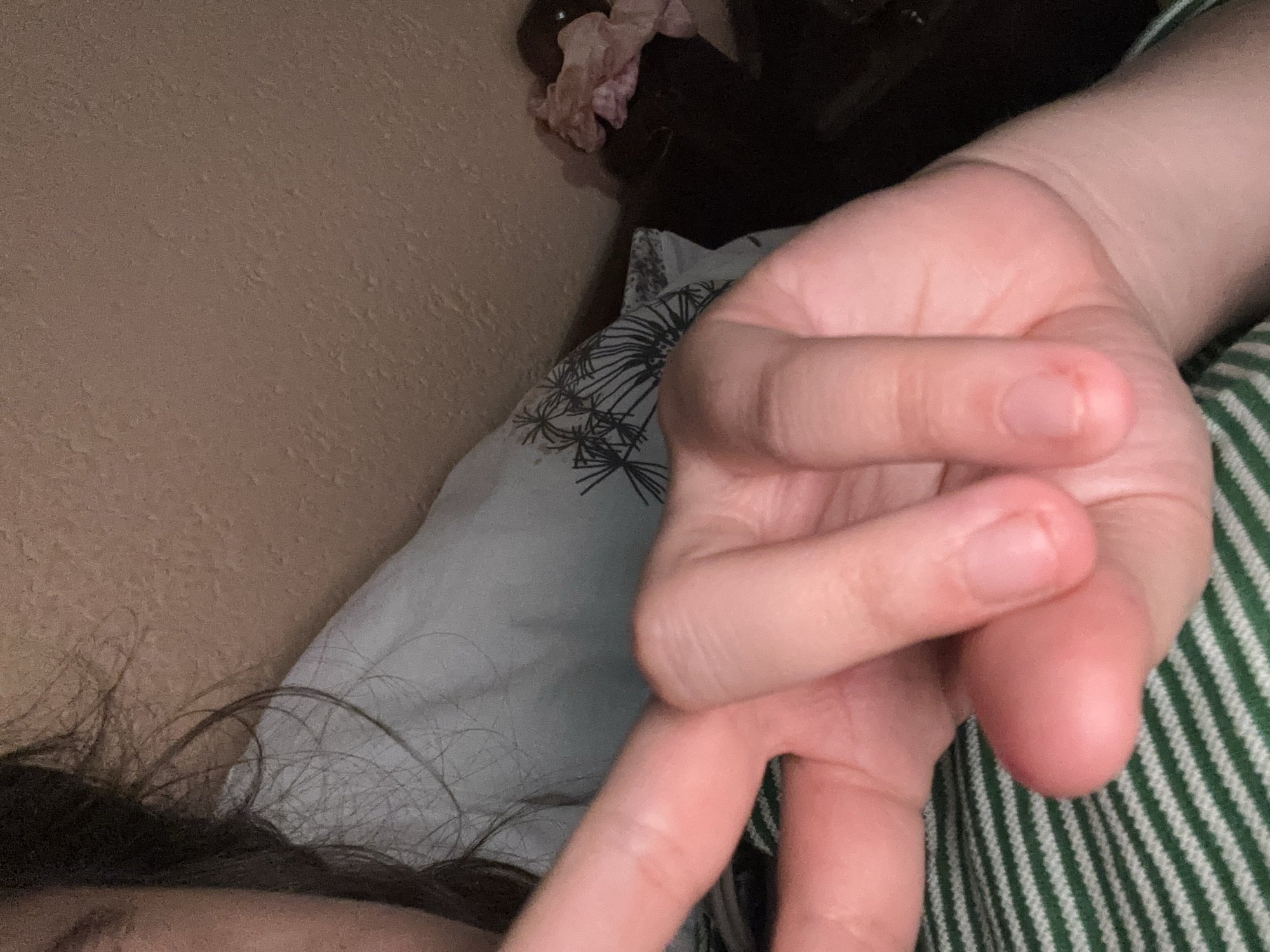 A close-up indoor shot shows a fair-skinned hand making a peace sign. The hand is positioned over a patterned pillow and a green-and-white striped fabric. Part of a person's face and dark hair are visible in the lower left corner. The background includes a beige wall and some dark objects. The location, Garmen, Bulgaria, is not discernible from the visual information.
