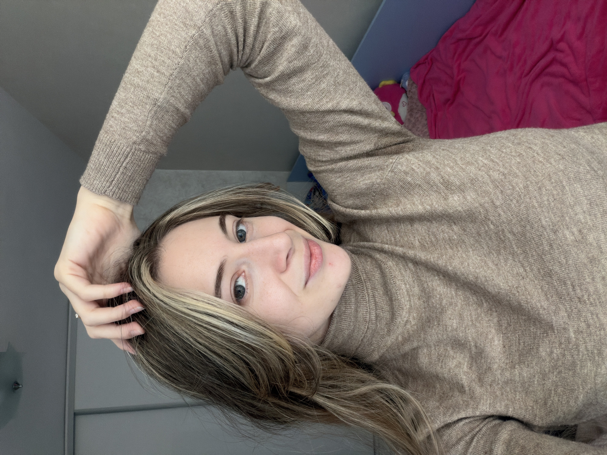 A woman is posing for a selfie. She is fair-skinned with brown hair that has blonde highlights. She is wearing a tan turtleneck sweater. Her left arm is raised with her hand near her head. The background includes a grey wall and a blue shelf with a pink blanket on it. The location cannot be determined from the image.