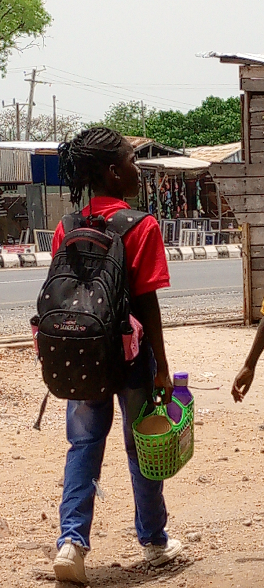 A person walks along a sandy, dusty road in Adankolo, Nigeria. The person, facing away from the camera, has braided hair pulled back in a bun. They are wearing a red shirt and blue jeans with a rip at the knee. A black backpack with white polka dots is slung over their shoulders. In their right hand, they carry a green plastic basket containing a brown coconut and a purple liquid bottle. The scene includes a roadside with a paved street and commercial buildings constructed from metal or wood, suggesting an urban setting. Power lines are visible in the background. Another person's arm is visible on the right side of the image.