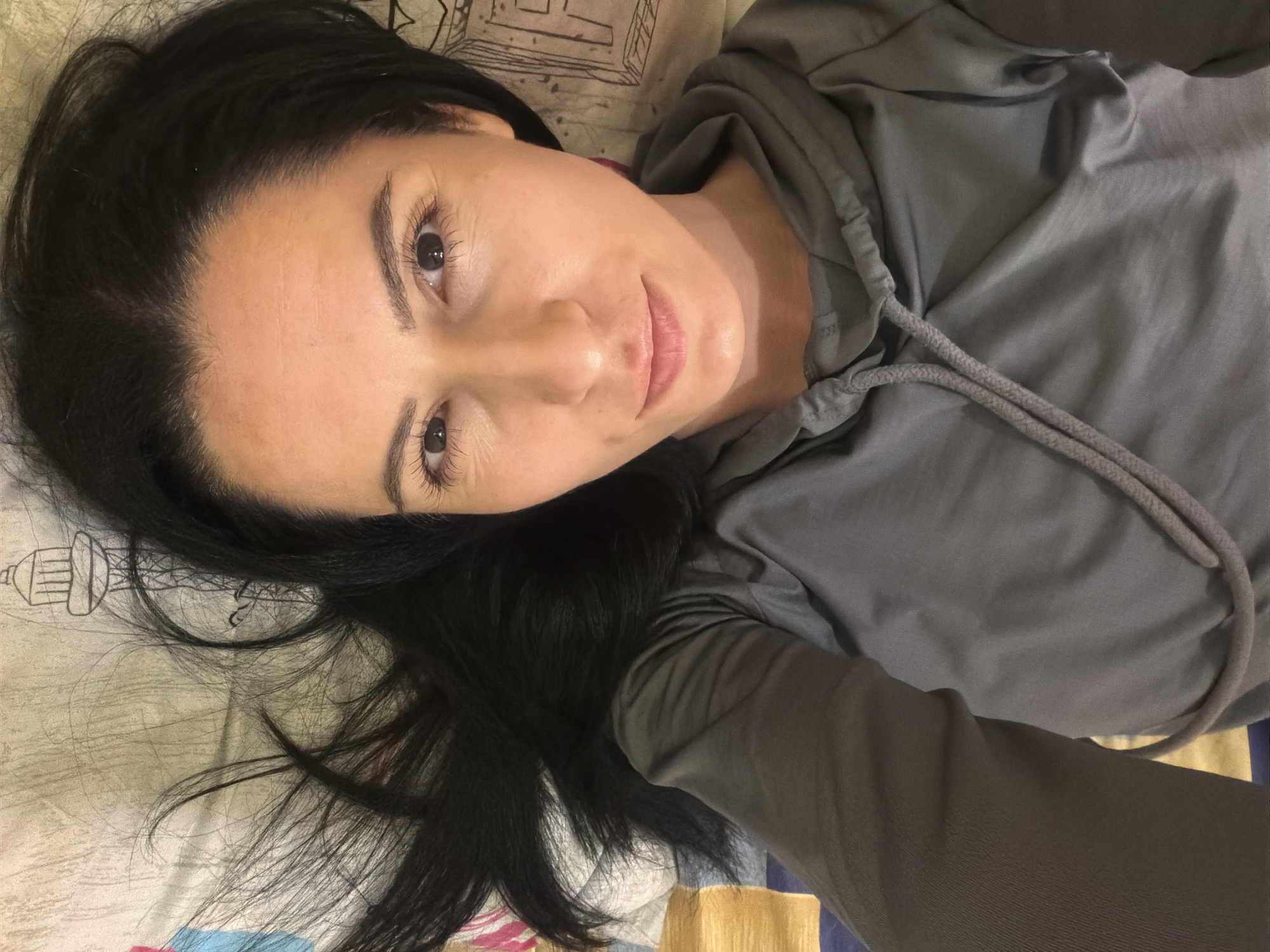 An overhead photograph depicts an adult female lying supine on a patterned bed surface. The subject has dark, shoulder-length hair fanned out around her head and a fair complexion. She is wearing a grey, long-sleeved hooded sweatshirt with visible drawstrings around the neck. Her eyes are open and directed towards the camera, and her mouth is closed with a neutral expression. The background consists of a light-colored sheet or duvet cover featuring various illustrations, including a prominent black outline drawing of a water tower with a domed top on a lattice base, located to the right of the subject's head. This illustration strongly resembles the Pleven Water Tower, indicating the scene is situated within a residence or accommodation in Pleven, Bulgaria. Additional patterns on the bedding include sections of blue, yellow, and red geometric shapes.