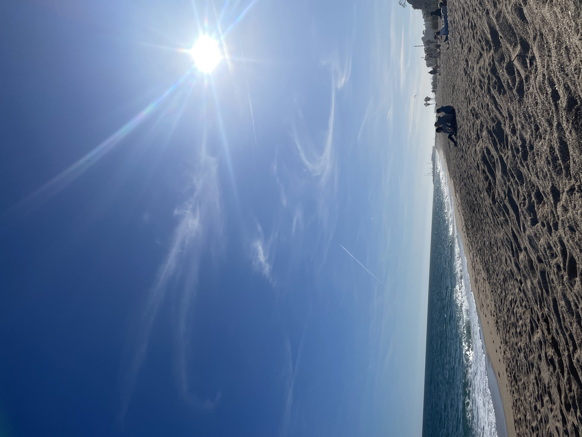 Here's a detailed description of the scene:
The image captures a bright, sunny day at the beach in Canet De Mar, Spain. The sky is a vivid blue, with wispy cirrus clouds and a visible contrail from an airplane. The sun shines intensely, creating lens flare effects.
The Mediterranean Sea is visible, with gentle waves lapping against the shore. The beach is composed of dark sand, marked with textures and patterns. Several people are scattered along the shoreline, engaging in activities like sunbathing and sitting in groups.
Buildings and other structures are visible along the horizon in the background, providing a sense of the town's location. The overall composition suggests a typical sunny day at a popular beach location.