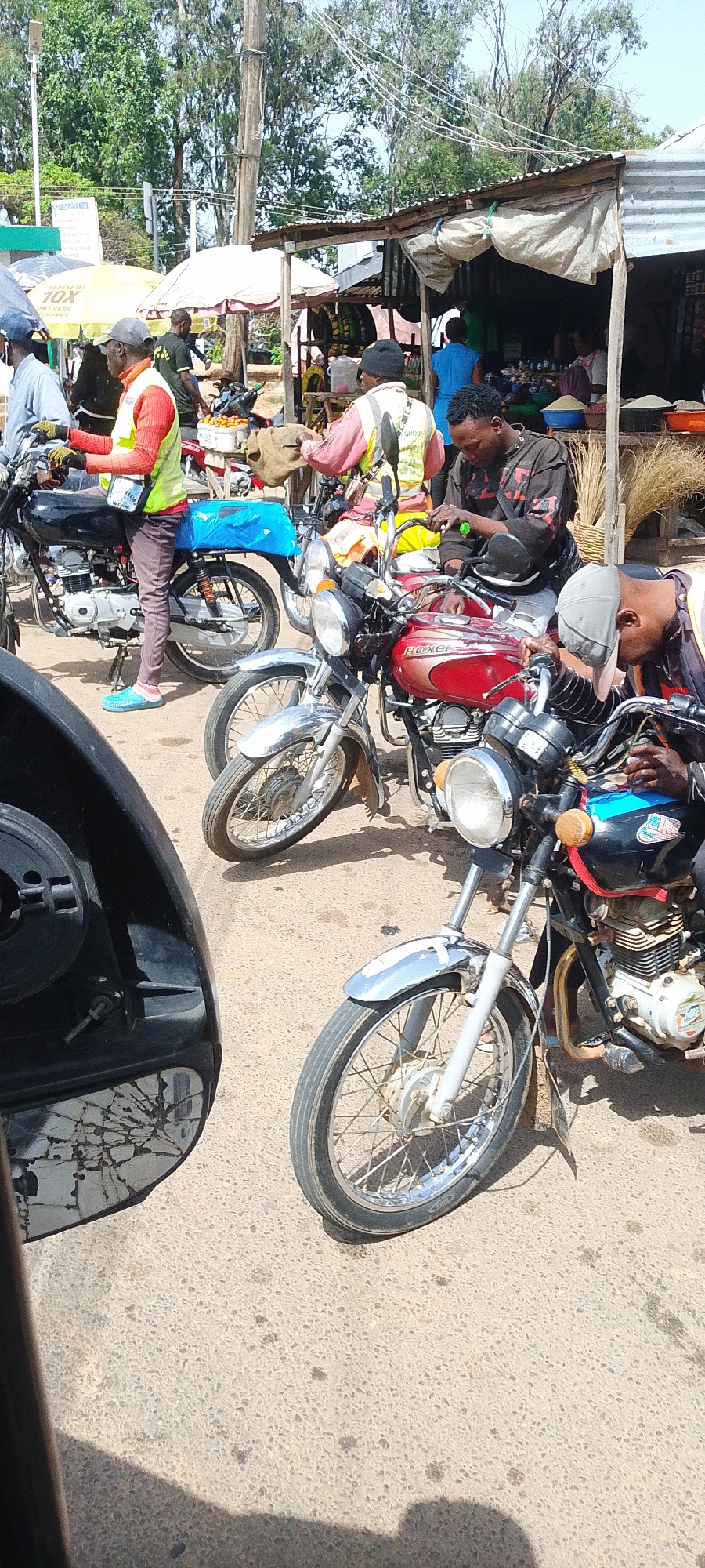 This media file captures a bustling street scene in Bukuru, Nigeria, likely during the daytime with clear weather. The foreground is dominated by several motorcycles, suggesting a mode of transportation and potentially a taxi or delivery service area. In the mid-ground, various individuals are engaged in activities related to commerce. One person, wearing a red and yellow vest, stands near a motorcycle. Another individual in a similar vest is seen interacting with a sack. Several market stalls are visible, featuring goods like rice and what appear to be fruits, with some vendors or customers in the background. A prominent red and silver motorcycle has "BOXER" written on its side. The overall atmosphere appears to be that of a busy market or transport hub. The presence of a cracked mirror in the lower left suggests the perspective is from inside a vehicle.