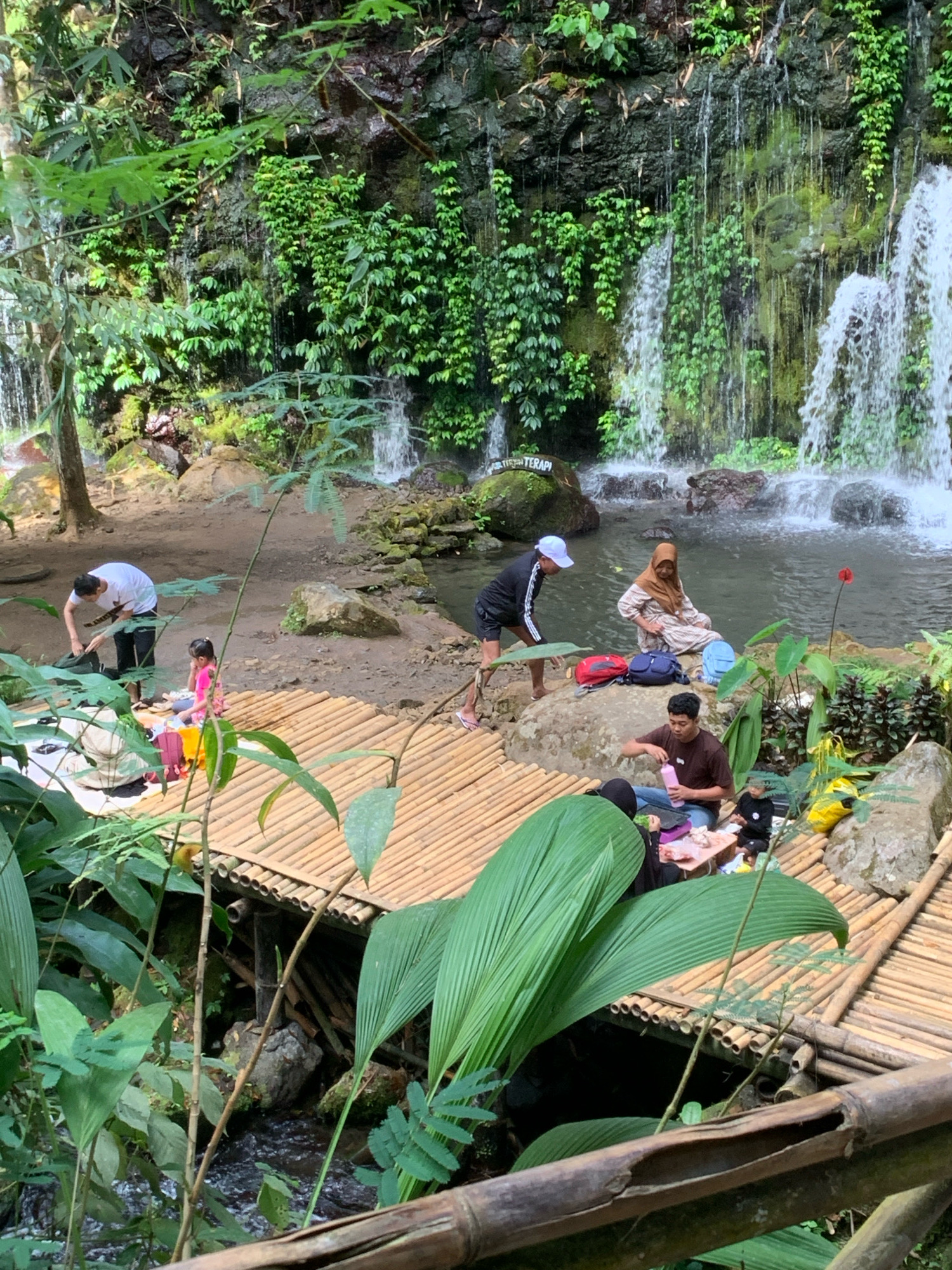 The image depicts a serene natural setting near a waterfall, likely in Pronojiwo, Indonesia. A group of people are engaged in a picnic or relaxation activity on a bamboo platform.
In the foreground, a bamboo platform is partially obscured by lush green foliage. On the platform, two individuals are seated. One, wearing a black hijab and dark clothing, is focused on food laid out on a mat. Another person, wearing a brown t-shirt and jeans, is holding a pink water bottle. A child in dark clothing is also present on the platform.
Further back, on a sandy area adjacent to a pool of water, a man in a white t-shirt and dark pants is packing items into a bag. A young girl in a bright pink outfit is seated nearby. To the right of the water, a woman wearing a hijab and a patterned dress is sitting on a rock, with several backpacks at her side. Another person in athletic wear and a white cap is standing near the edge of the water.
The environment is dominated by a rocky cliff face from which multiple streams of water cascade down, forming a picturesque waterfall. The area is rich with vegetation, including various types of green plants and mosses clinging to the rocks. The water flows into a calm pool. In the background, a sign with text, partially visible, reads "TERAPI". The overall impression is one of a tranquil natural retreat.
