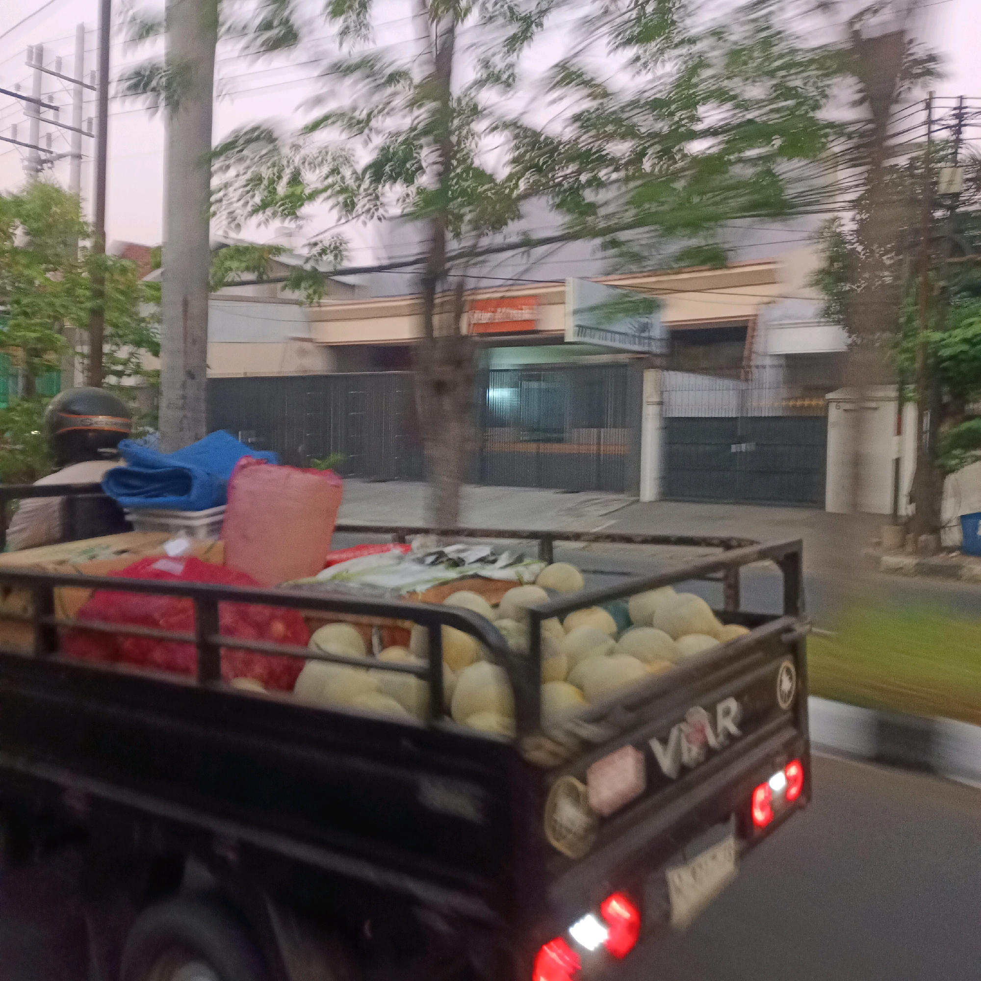 The image, captured in Surabaya, Indonesia, shows a dark-colored pickup truck in motion, indicating it is driving on a road. The truck bed is loaded with produce, primarily what appear to be melons, and other bundled items, including a large pink sack and a blue tarp. A motorcycle helmet is visible on the back of the truck, behind the driver's seat. The background features a blurred street scene with buildings, trees, and utility poles, suggesting an urban environment. The motion blur indicates the photograph was taken from another moving vehicle.