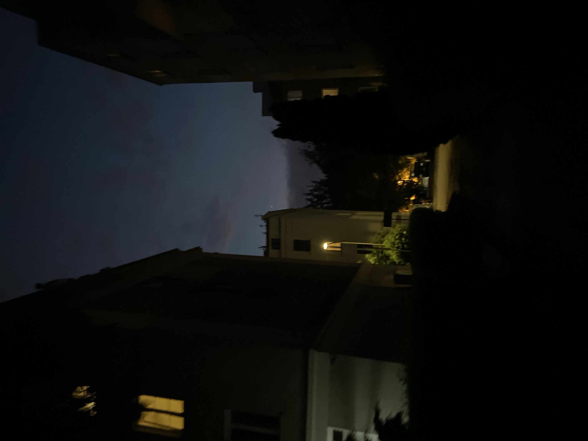 The image depicts an urban scene at dusk or night, likely in Warsaw, Poland, characterized by low light conditions and architectural structures. On the left, a building facade is visible, with illuminated windows casting a yellow glow. To the right and further in the background, another building with similarly lit windows is partially obscured by darkness. In the center of the frame, a street or pathway is visible, leading towards more illuminated buildings and trees. A street lamp is also present, providing additional light. The sky is a deep blue, suggesting the transition from evening to night. The overall impression is one of a quiet, dimly lit urban environment.