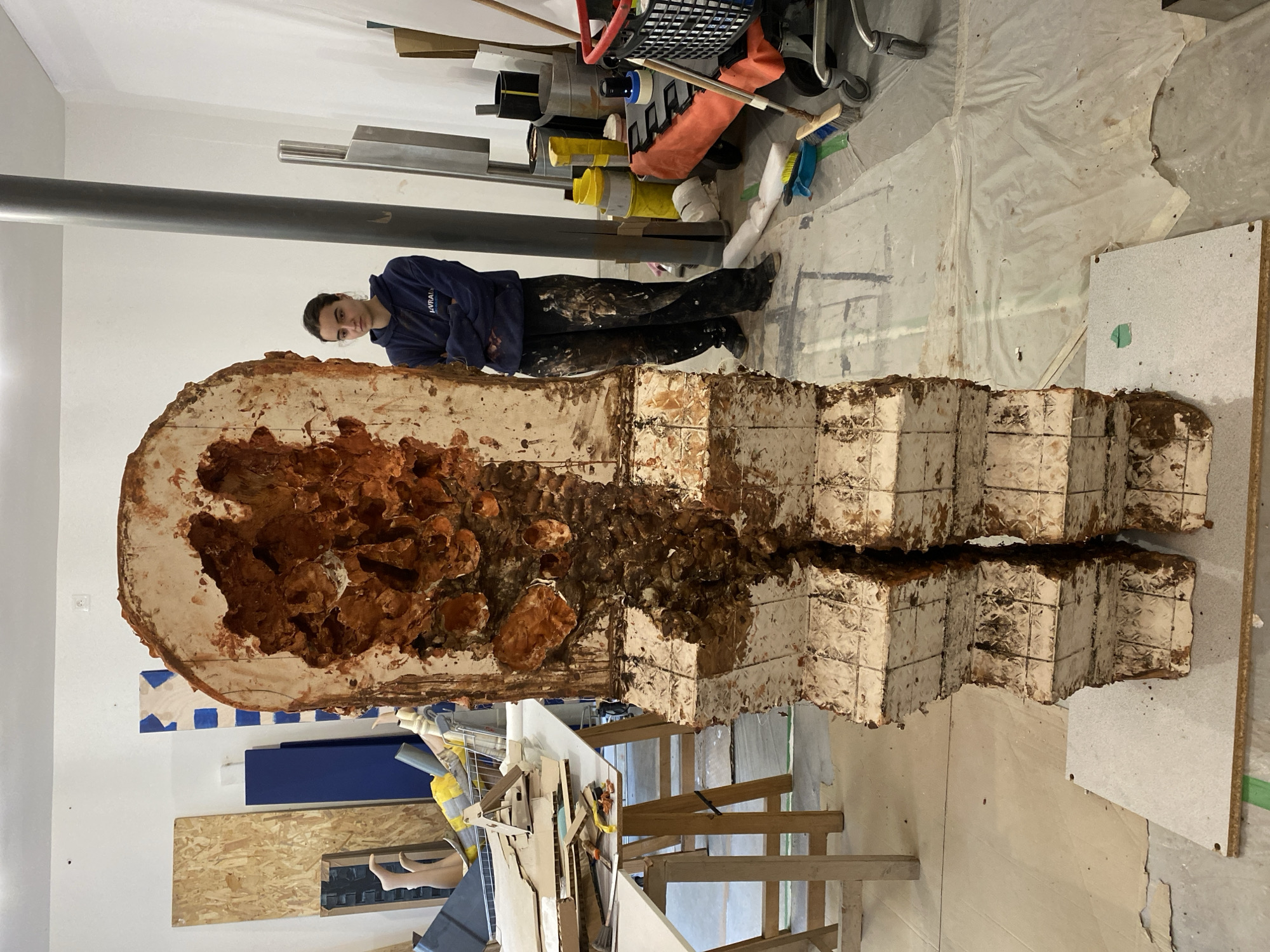 The image shows an indoor scene that appears to be an artist's workshop or studio. A person with dark hair and a blue sweatshirt is standing next to a large, abstract sculpture made of stacked, textured blocks with a hollow, reddish-brown interior. The sculpture is split into two sections. Various materials and tools are scattered around the space, including paint cans, metal bars, wooden planks, and a metal shelving unit. The floor is covered with a plastic sheet, indicating ongoing or recent art-making activity. It is difficult to deduce the exact location in Paris, France, as there are no specific identifying landmarks or street signs visible in the image.