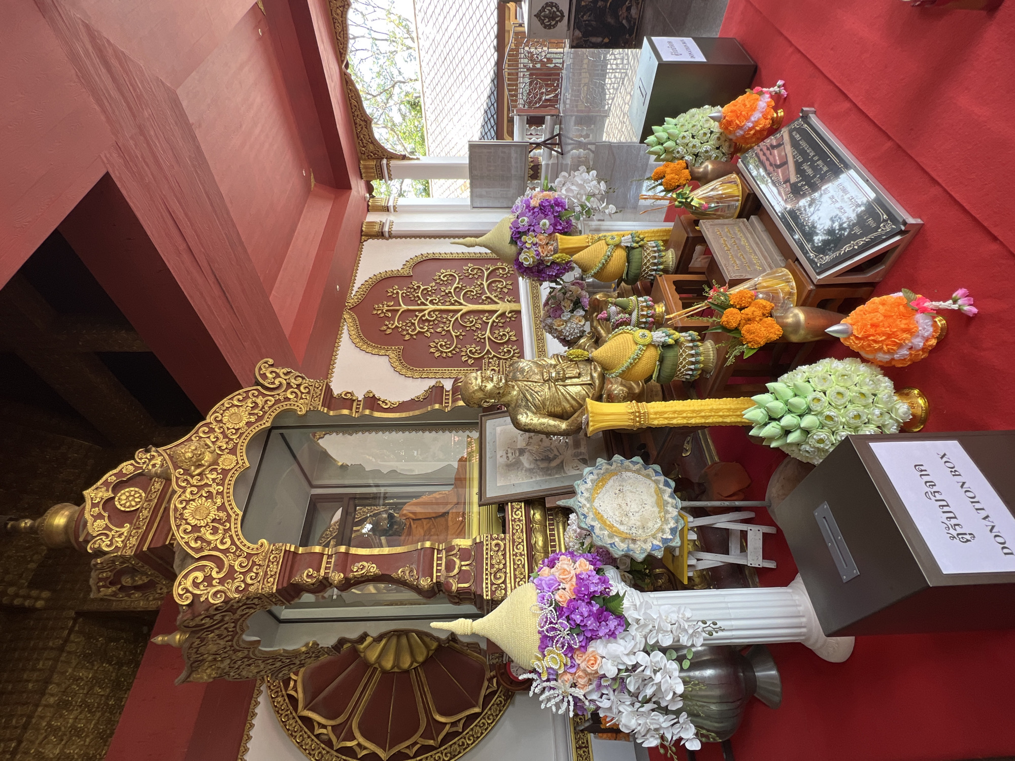 The image depicts a shrine area, likely within a temple or Buddhist site, in Baan Hua Thanon, Thailand. The scene is ornate, with abundant use of gold and vibrant colors. At the center of the frame, a glass display case houses a seated monk figure dressed in saffron robes. Adjacent to this display are several golden Buddha statues, a framed photograph of a monk, and an assortment of offerings.
The offerings include large floral arrangements in various colors such as white, purple, pink, and orange, along with sculpted lotus flowers made from green and white material. Tall yellow candle-like structures and incense sticks are also visible among the displays. Several decorative conical structures, possibly representing stupas or traditional ceremonial items, are placed on stands.
In the foreground, a dark grey donation box with a slot on top is prominently placed, featuring Thai script and the English text "DONATION BOX". To the right, another, larger green donation box is visible. A reflective surface, likely a polished floor, mirrors the elements of the shrine. In the background, through an open archway, a glimpse of the exterior can be seen, with a building roof and trees. The architectural style of the shrine itself is elaborate, featuring intricate gold carvings and a red ceiling. The overall impression is one of reverence and decoration associated with religious practices.