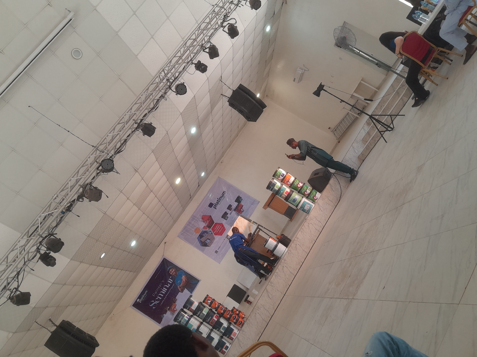 The scene is an indoor seminar event, likely a conference or workshop, in an unknown location. Overhead, a metal truss system supports numerous spotlights and black speakers. Banners promoting "Platinum Painter's Seminar" and "Platinum" paint products are displayed on the walls. A presenter wearing a blue shirt is arranging paint cans on a table. An audience member is capturing the scene with a mobile device. A standing fan is visible in the background. Audience members are seated in red chairs. The floor has a marble-like tile pattern.