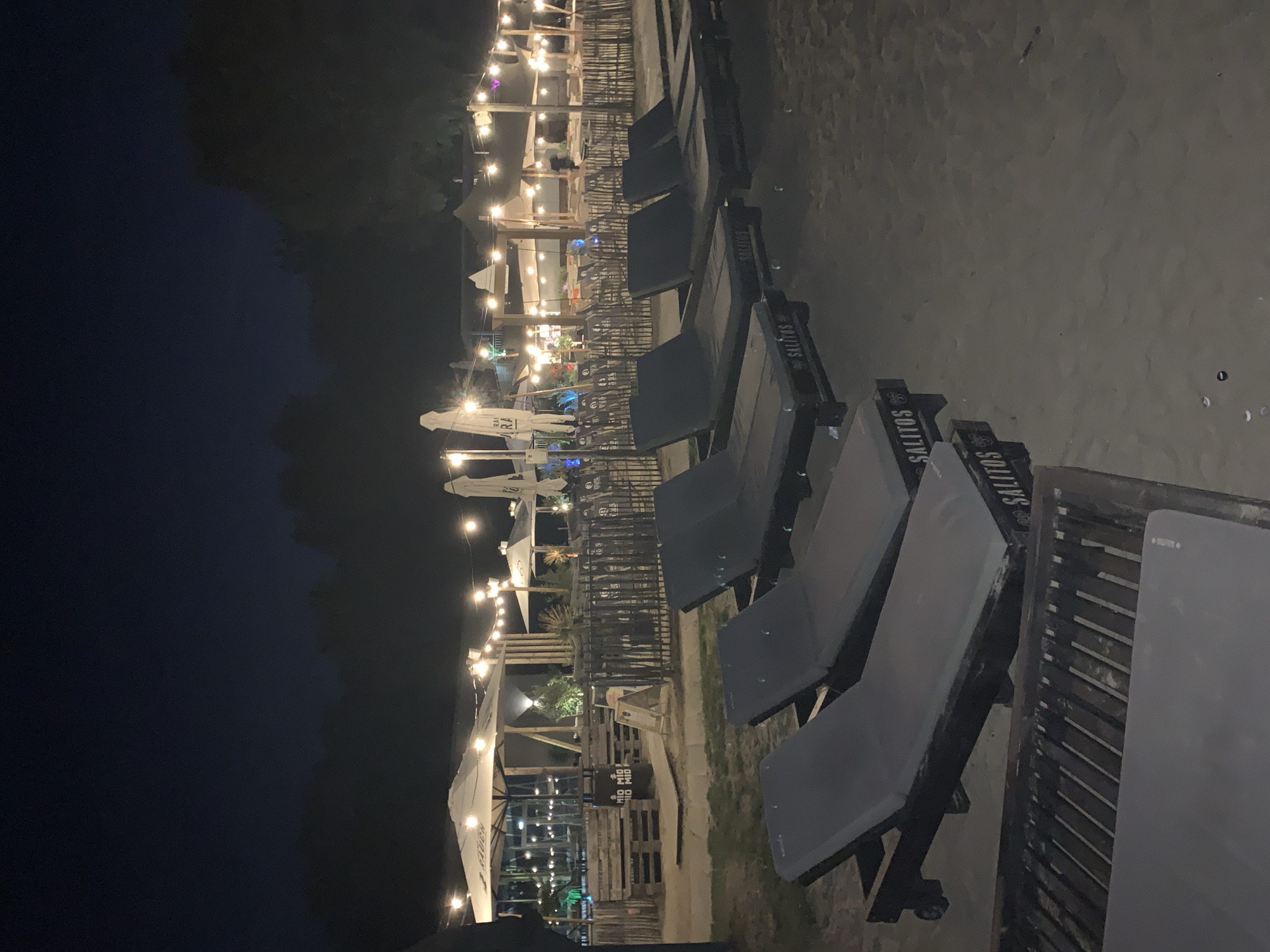The image depicts a beachside seating area at night in Rotenburg, Germany. Several empty, dark gray lounge chairs are arranged in rows on the sand, with the foreground occupied by two lounge chairs, one slightly behind the other. These chairs have wooden frames with "SALITOS" branding visible on the sides. Further back, a row of additional lounge chairs is present.
Behind the lounge chairs, a fenced-off area contains multiple tables and chairs, presumably for dining or relaxation. White umbrellas are set up over some of these tables. String lights are strung across poles, illuminating the area. In the background, trees form a dark silhouette against the night sky. The overall atmosphere suggests a casual, outdoor venue open after dark.
