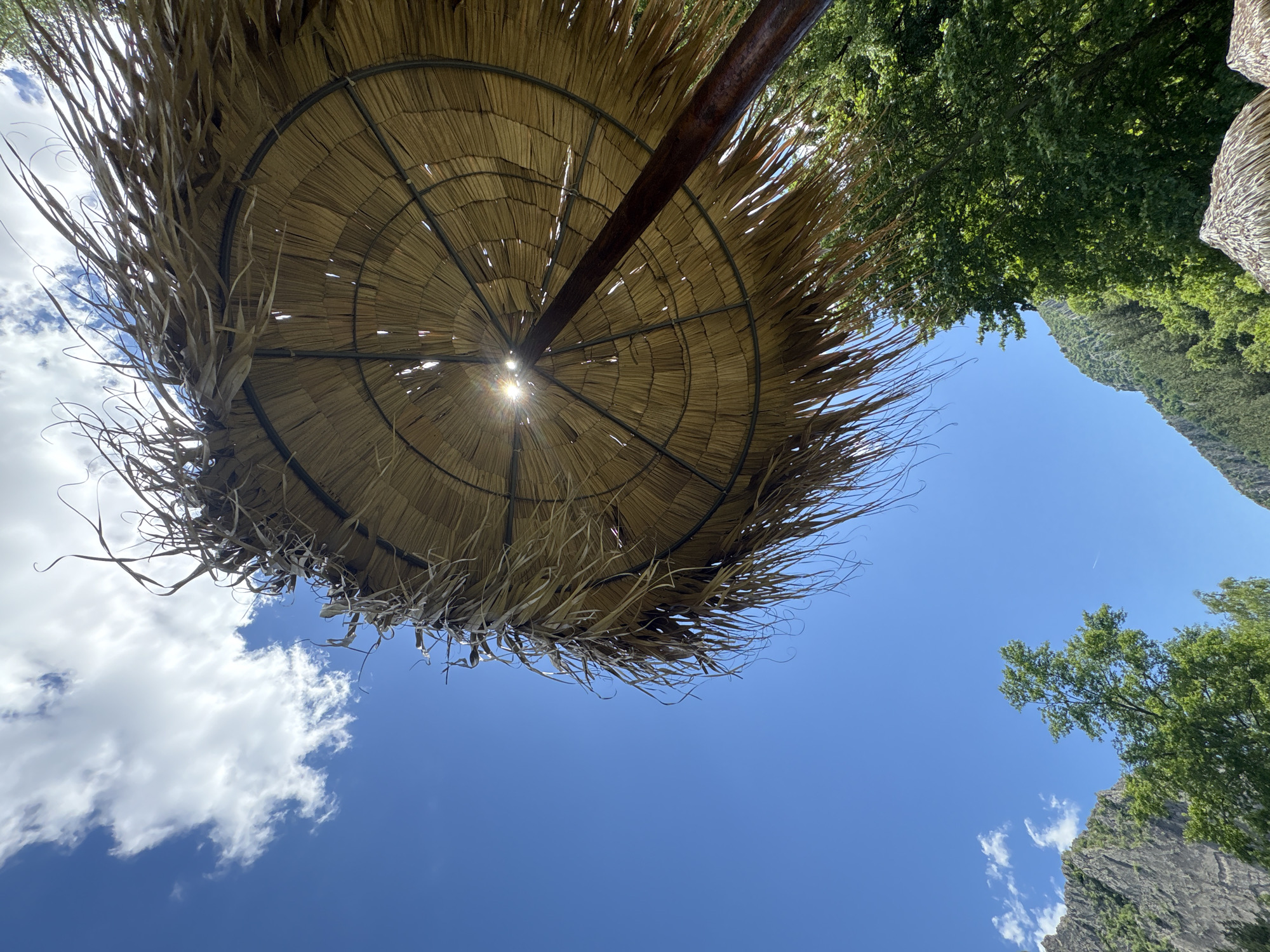 An upward-angle shot reveals a large, thatched umbrella casting a shadow under a bright blue sky with scattered white clouds. The umbrella's straw material is worn and frayed, with strands extending outwards. Sunlight pierces through gaps in the thatch, creating bright spots. A thick, dark brown pole supports the umbrella, angled upwards. In the background, lush green trees and a rugged, grey mountain range are visible. The scene suggests a natural, possibly recreational area.