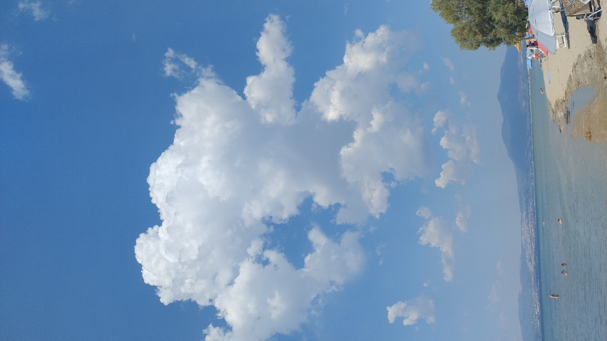 A wide-angle, vertical shot captures a bright, sunny day at a beach. The sky is a vibrant blue, dotted with fluffy white cumulus clouds. In the foreground, a large cloud dominates the upper left portion of the frame. To the right, the scene transitions to a sandy beach with a few people lounging on chairs under umbrellas. Beyond the beach, the clear, blue sea stretches to the horizon, meeting a distant, mountainous coastline with a town nestled at its base. Several people are visible in the water, some swimming or wading. A lone tree stands on the edge of the beach, providing shade. The overall mood is one of leisure and relaxation on a beautiful summer day. The location appears to be Neoi Epivates, Greece, suggested by the mountainous coastal scenery and the presence of a beach resort. No visible text is present in the image.