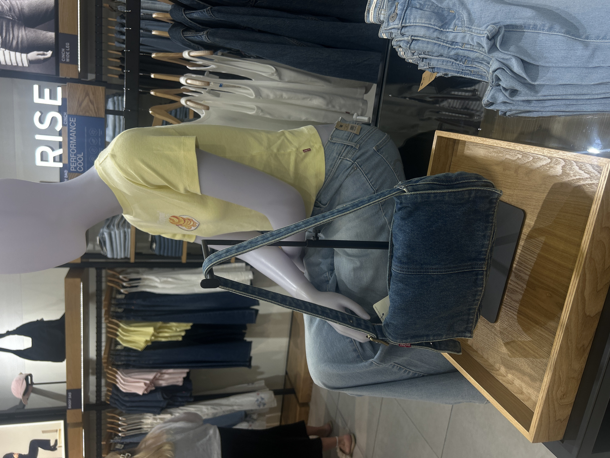 The image shows a mannequin inside a clothing store, likely in Patong, Thailand. The mannequin is wearing a yellow shirt and light-wash jeans. A denim purse is placed next to the mannequin inside a wooden tray. Racks of clothing are visible in the background, including tank tops, jeans, t-shirts, and blouses. A sign advertising "Rise Performance Cool" clothing is visible to the left of the mannequin.