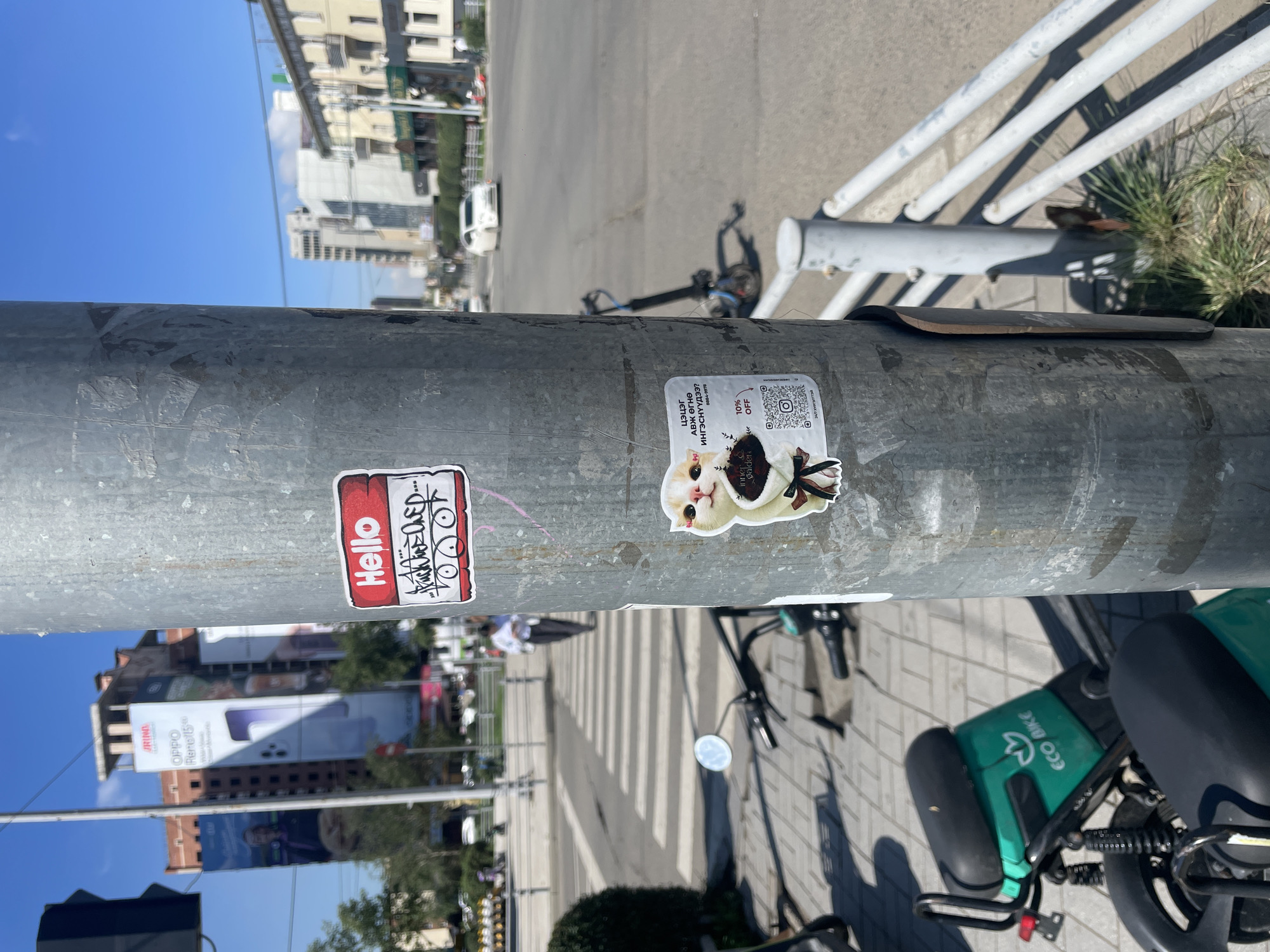In Ulaanbaatar, Mongolia, on a sunny day, a close-up shot captures a section of a metal lamppost adorned with two stickers. The lamppost is positioned on a sidewalk next to a street where traffic is visible. In the background, large billboards are affixed to buildings, advertising products. One billboard prominently displays an advertisement for "OPPO Reno 15."
The top sticker on the lamppost is a red and white nametag-style sticker with the word "Hello" written in red. Below it, handwritten graffiti in black ink is present.
The bottom sticker features a cartoon illustration of a cat with white and orange fur, holding a black flower with a white ribbon. Text in Mongolian script is visible on this sticker, along with a QR code and a "10% OFF" promotion.
To the left of the lamppost, an "Eco Bike" electric scooter is parked on the sidewalk. A pedestrian is walking across a crosswalk in the mid-ground. The scene suggests an urban environment with commercial activity and public transportation options.