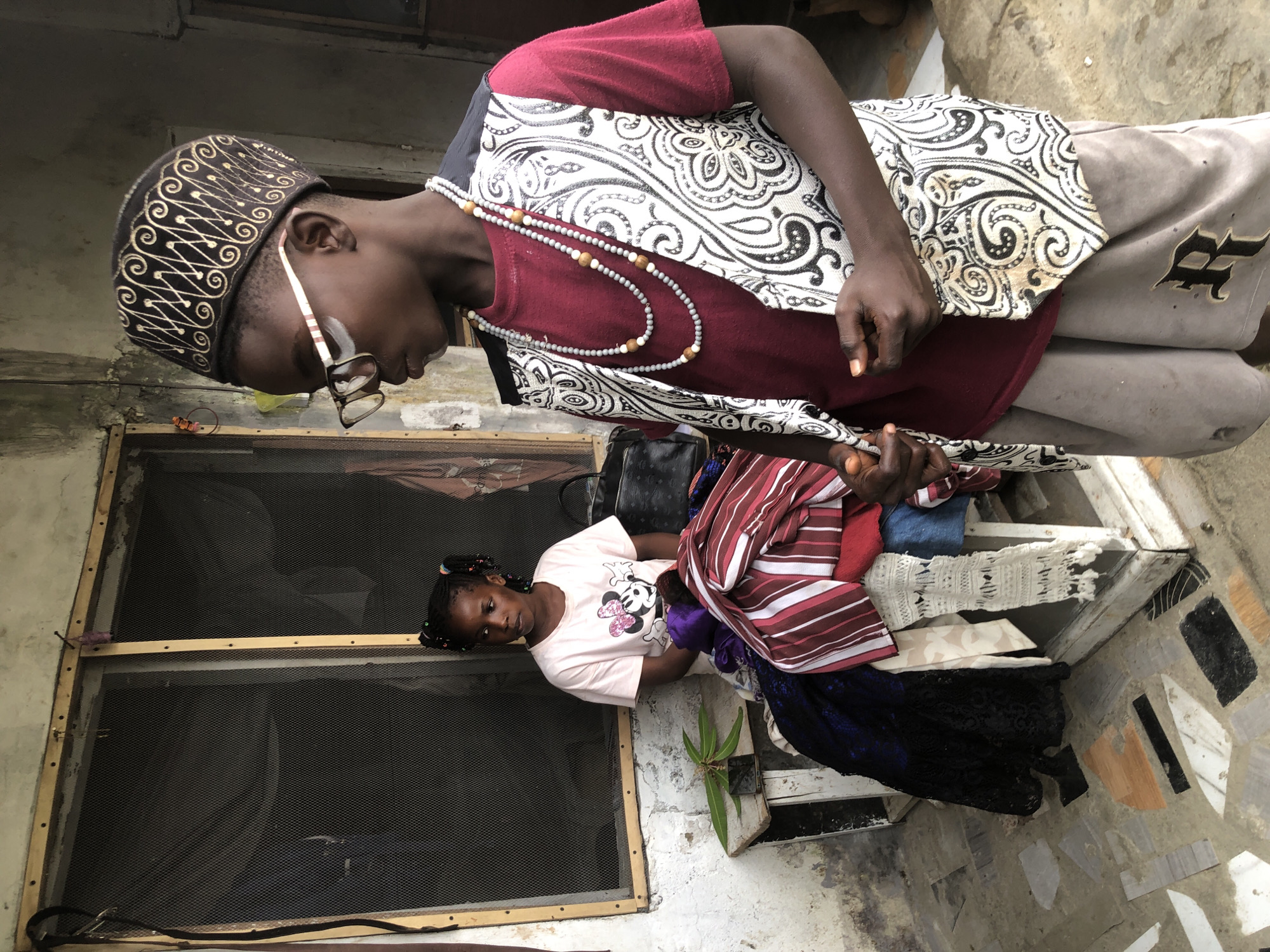 In Ado, Nigeria, an indoor scene captures two children. A boy, foreground right, wears a patterned cap, glasses, a burgundy t-shirt, a decorative vest with a black and white paisley pattern, and grey shorts. He is adorned with two beaded necklaces. To his left, a girl sits behind a collection of folded clothes. She wears a pink t-shirt with a Mickey Mouse graphic and has her hair in two braids. The children are positioned near a window with a screen. The floor is tiled with irregular pieces of stone.