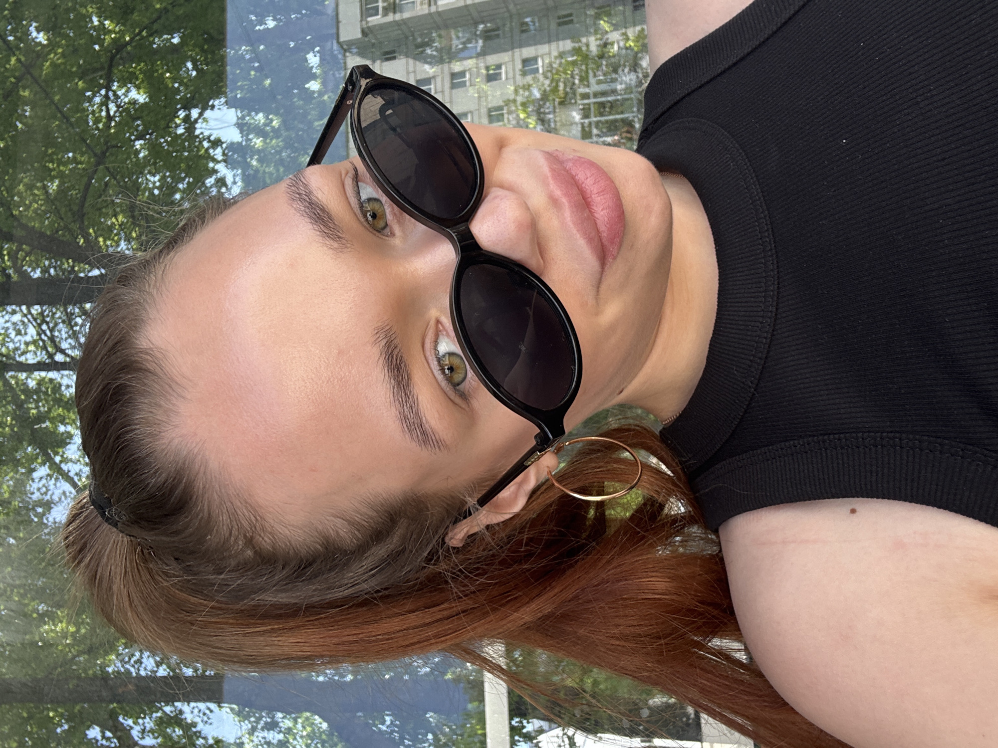 A woman with red hair and fair skin is shown in a rotated close-up selfie wearing black round sunglasses. She has green eyes and pink lips. She is wearing a black top, and a gold hoop earring is visible. The background appears to be a reflection of a city scene with trees and buildings. Based on the prompt, this is Chaikovsky, Russia.