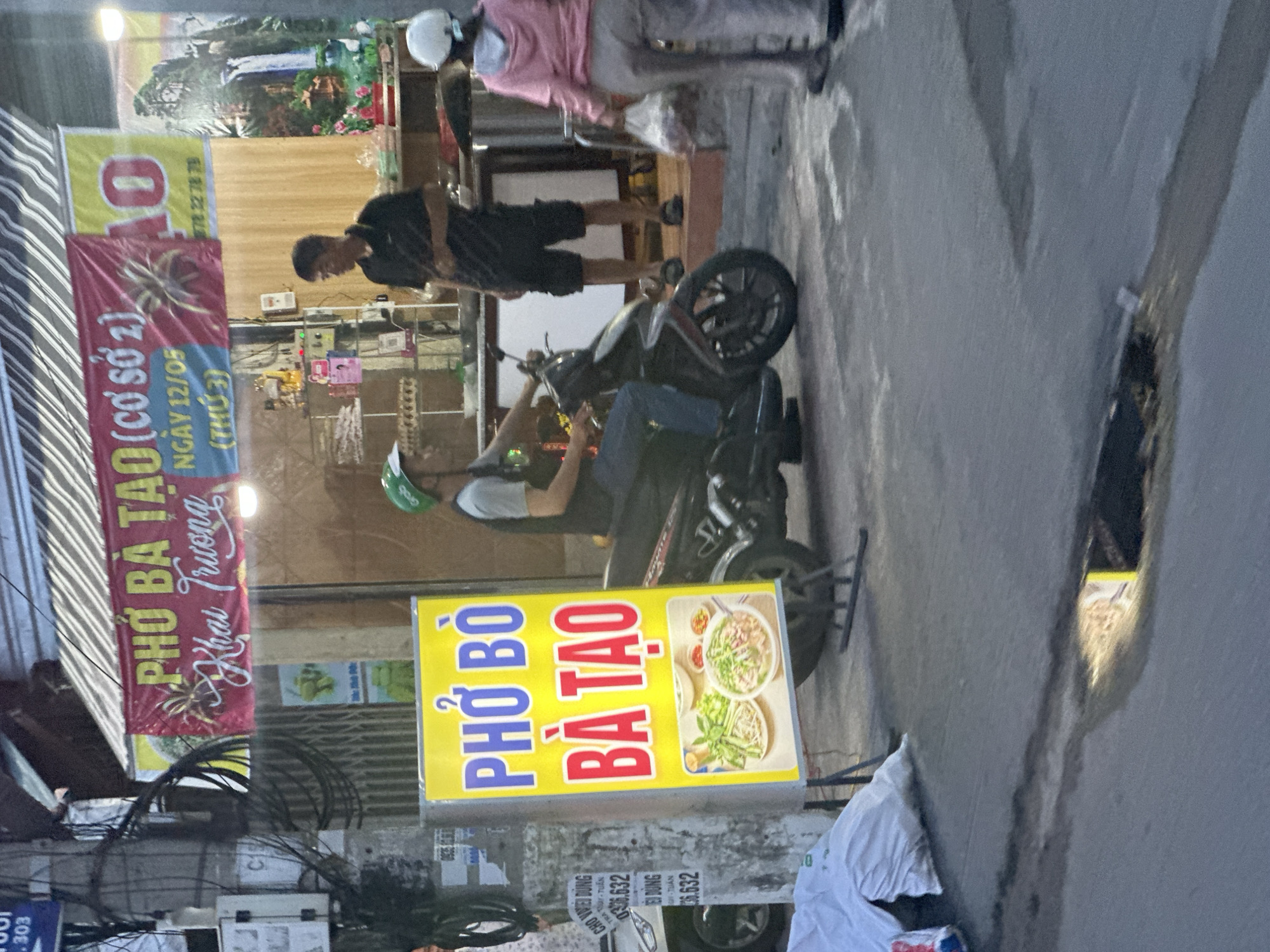 Here is a description of the image:
The image shows a street scene in Bac Nha Trang Ward, Vietnam. A Pho Bo Ba Tao restaurant sign is visible. A man is sitting on a scooter, and another man is standing nearby. A woman in a pink jacket is also visible in the background. The environment appears to be an urban street, with power lines and buildings visible.