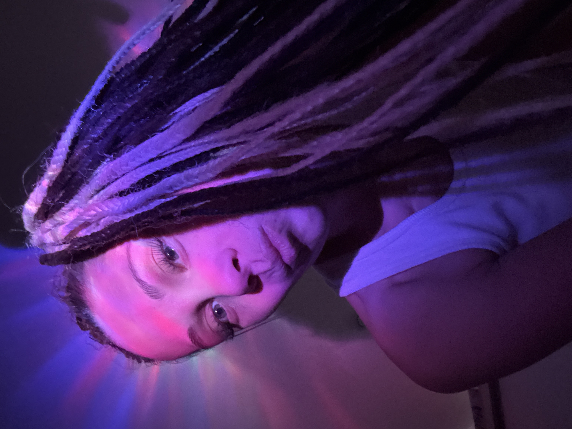 A close-up, dimly lit indoor photograph shows a person with black and purple synthetic dreadlocks, looking directly at the camera. The image is oriented as if the person is lying on their back or leaning back, with their head tilted. Their face is illuminated by vibrant artificial lighting in pink, purple, and blue hues, casting colorful patterns and glows on their skin and hair. The person has light-colored eyes and a neutral, slightly pensive expression, with their mouth slightly parted. They are wearing a white top, visible on the right side of the frame. The dreadlocks are prominent, showcasing their texture and varied dark and vivid colors. The background is dark and indistinct, indicating an indoor setting, likely in the evening or at night, given the reliance on artificial light sources. No specific objects or text are visible besides the subject. The setting is in Rusanovo, Russia.