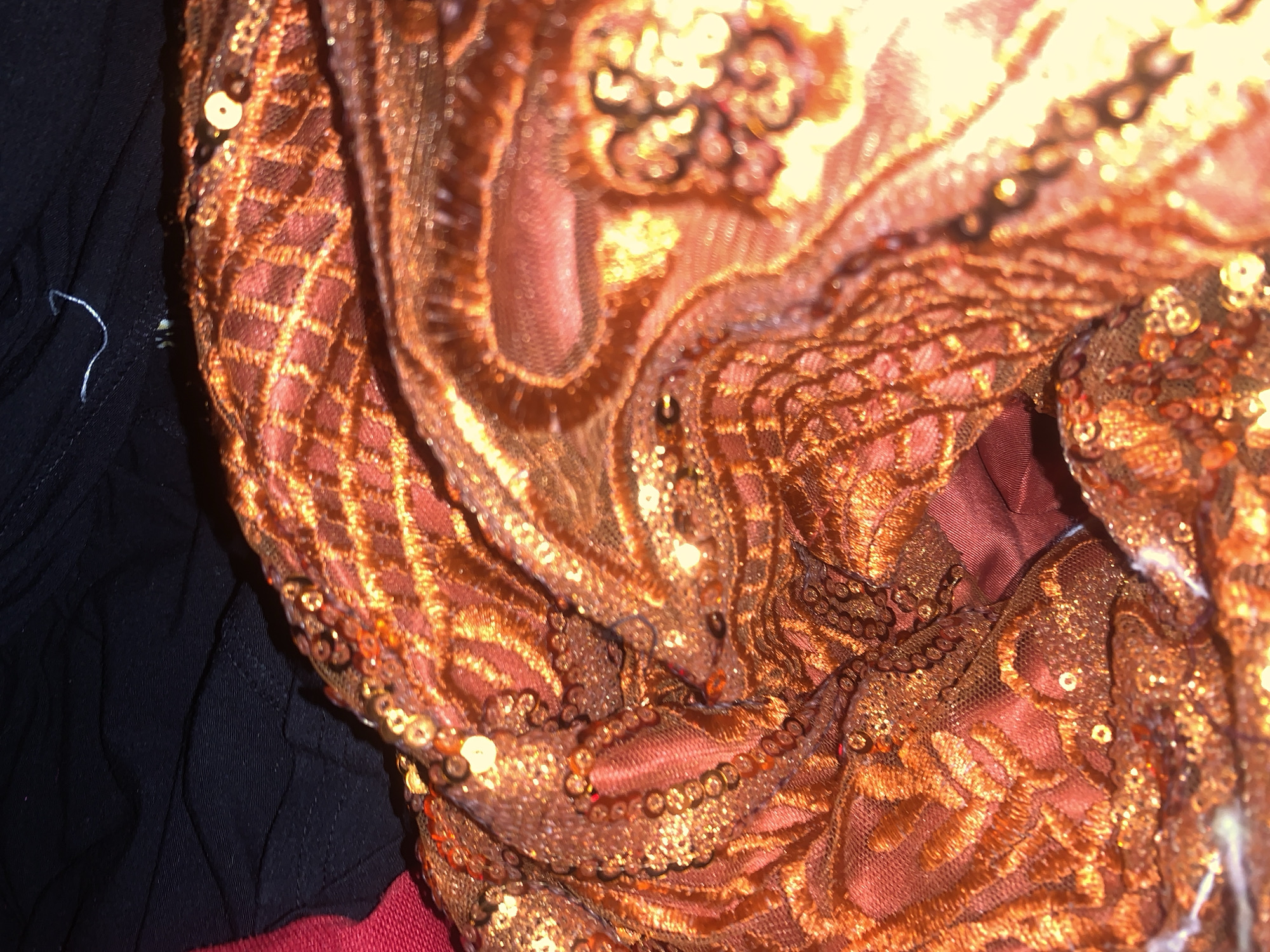 The image displays a close-up view of a draped, shimmering orange fabric heavily embellished with sequins and intricate embroidery. The fabric appears to be a sheer mesh overlaid with denser, textured patterns, and features small, circular sequins scattered throughout, reflecting light. Beneath the orange material, a darker, possibly red or maroon fabric is visible, suggesting a lining or another layer of clothing. To the right of the orange fabric, a plain black fabric is visible, also with some texture and possibly a stitching detail. The overall impression is one of opulent textile artistry. The image's context within Jalingo, Nigeria, is not visually discernible from the provided material.