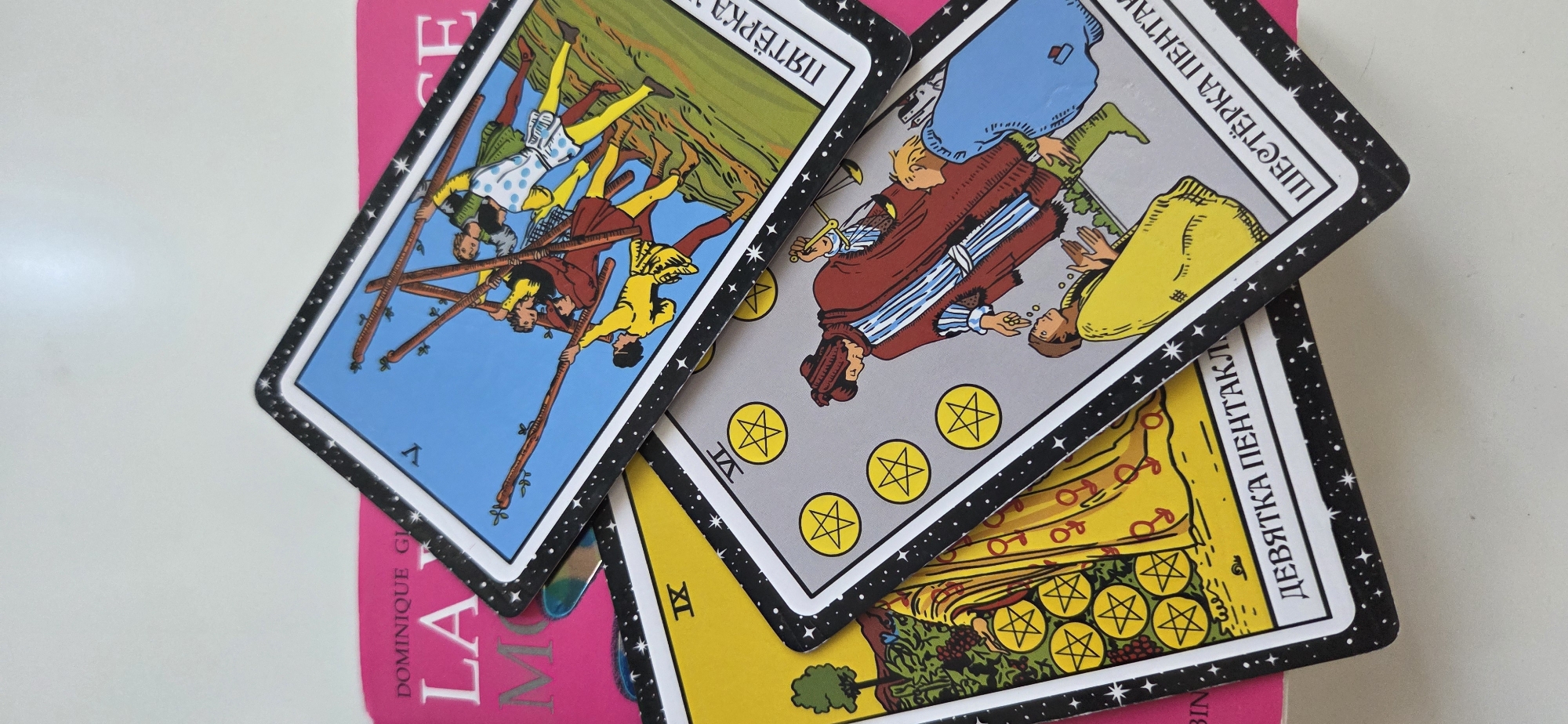 Three tarot cards are fanned out on a pink book. The cards are oriented diagonally, with their tops pointing towards the top left of the image. The top card, featuring a blue sky and a green hillside, depicts five figures struggling with wooden staffs. The middle card, with a grey background, shows two figures in historical attire. The bottom card, with a yellow background, displays a bounty of gold coins. The cards are edged with a black and white star pattern. The pink book beneath has visible text, including "DOMINIQUE GI" and "LA". The setting appears to be indoors, on a plain white surface. The lighting suggests daytime. The overall mood is neutral, with the focus on the tarot cards themselves.